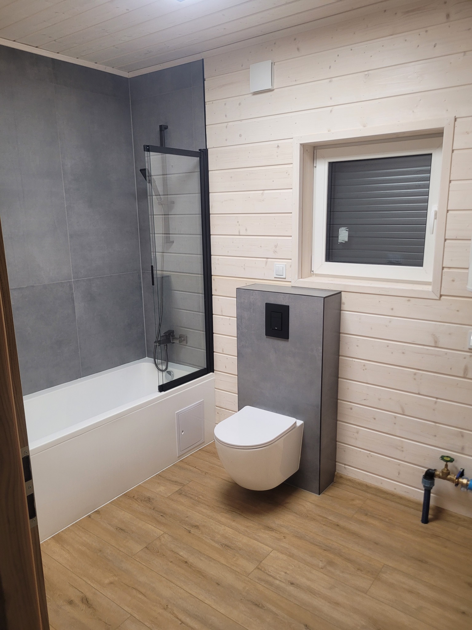Nowoczesna łazienka z drewnianymi panelami, szarą armaturą i podłogą imitującą drewno. Kabina prysznicowa z czarnymi ramami i wisząca toaleta.