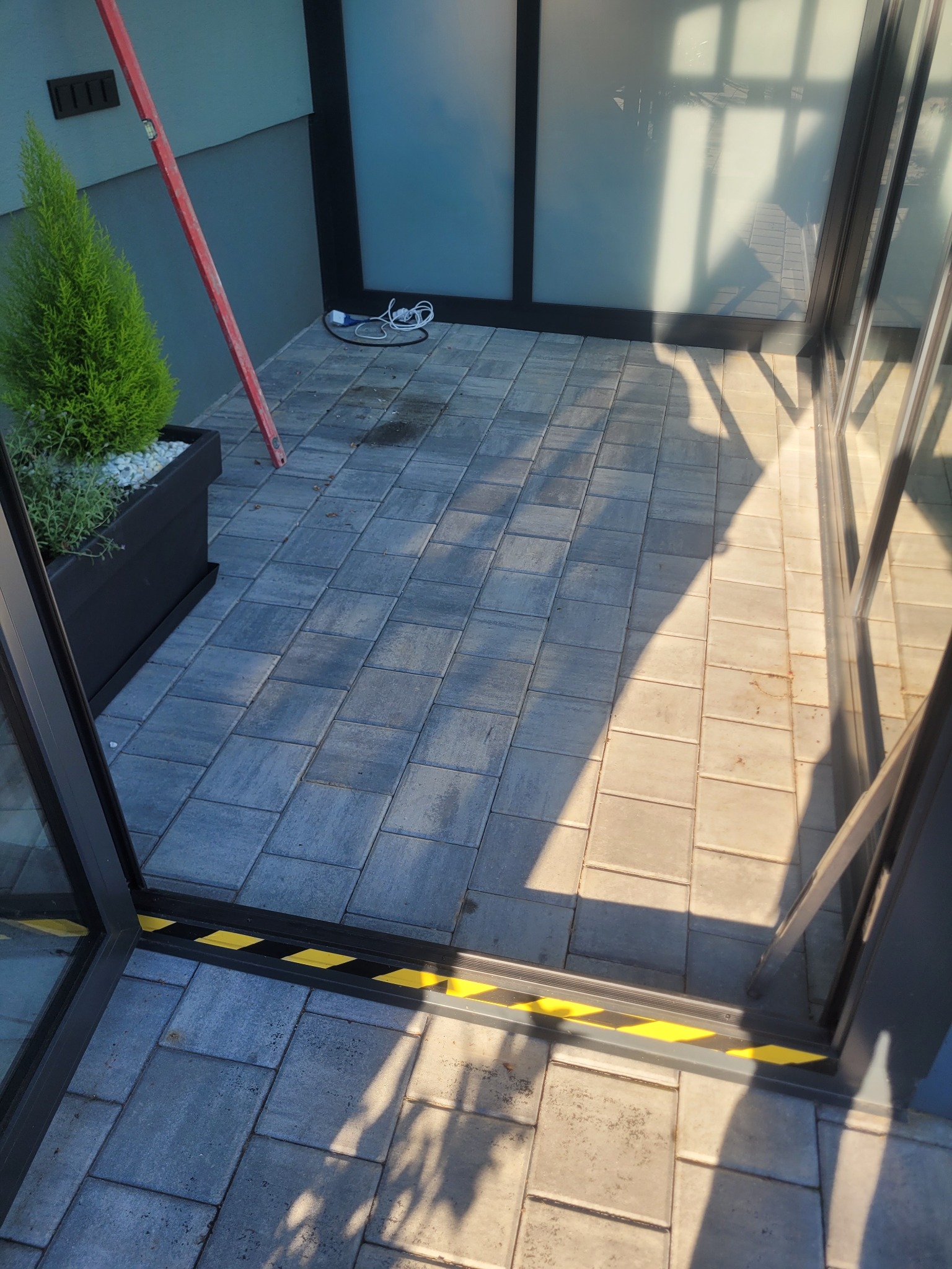 Ułożona kostka brukowa na tarasie z widocznym progiem oznaczonym taśmą ostrzegawczą. W tle roślina w donicy i pionowa poziomnica oparta o ścianę.