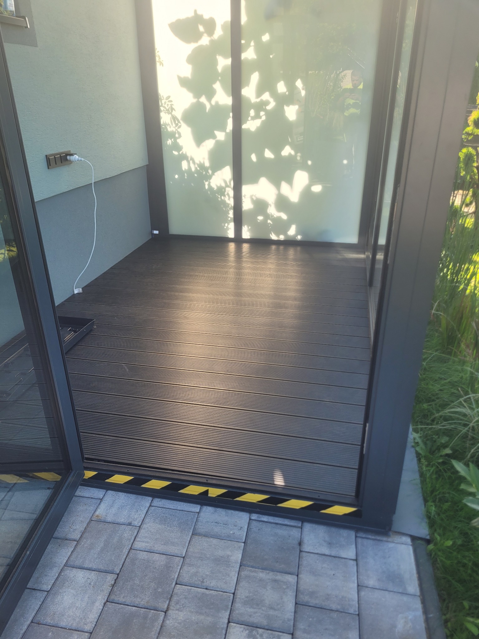 Zadaszony taras z desek kompozytowych, wykończony czarną listwą z żółtymi pasami. Widoczne szklane drzwi przesuwne i kostka brukowa. Świeże, jasne oświetlenie.