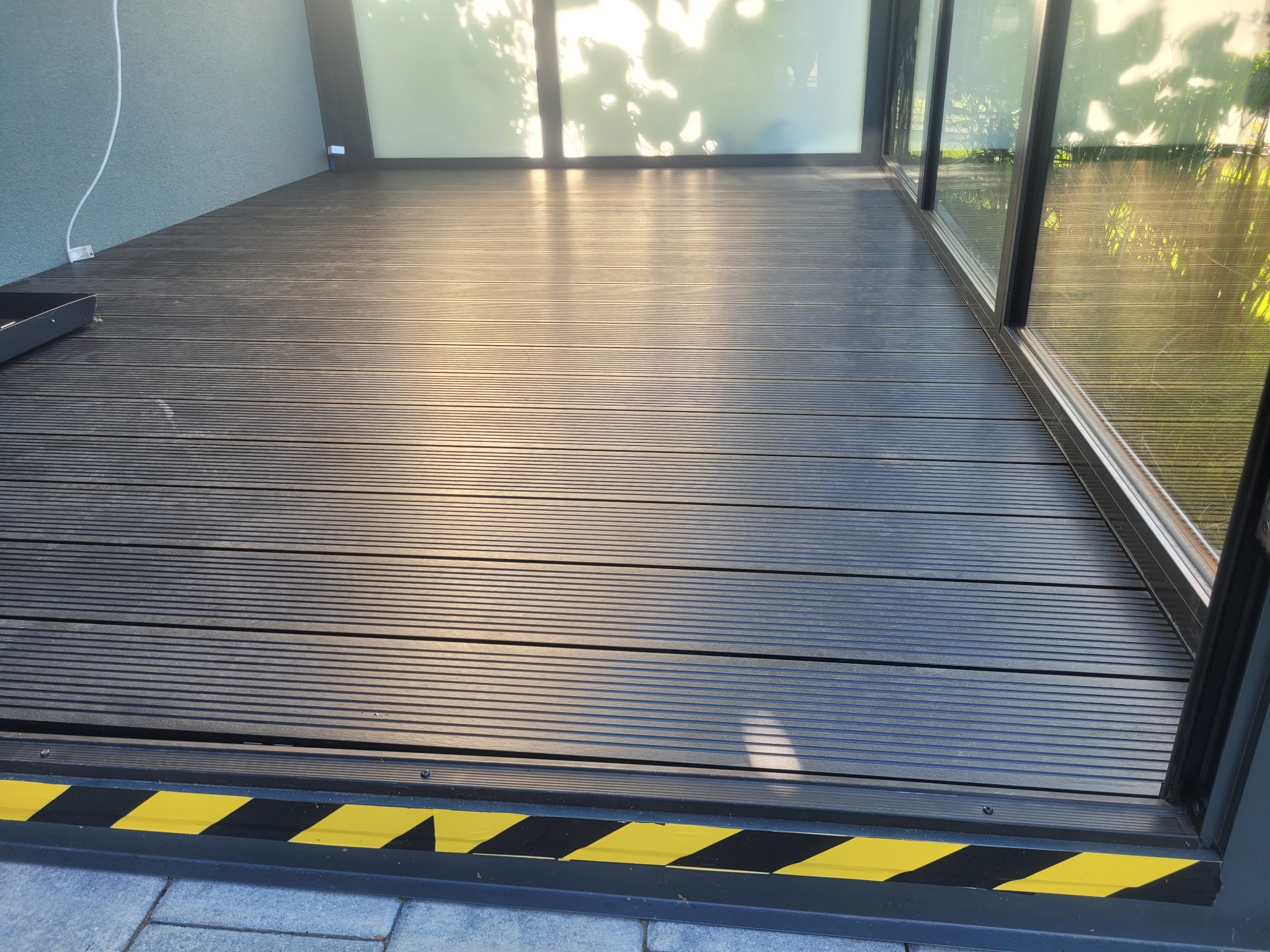 Wykończony taras z deski kompozytowej w Kamionnej, z żółto-czarnym pasem ostrzegawczym przy progu. Widoczne przesuwne okna tarasowe i fragment ściany.