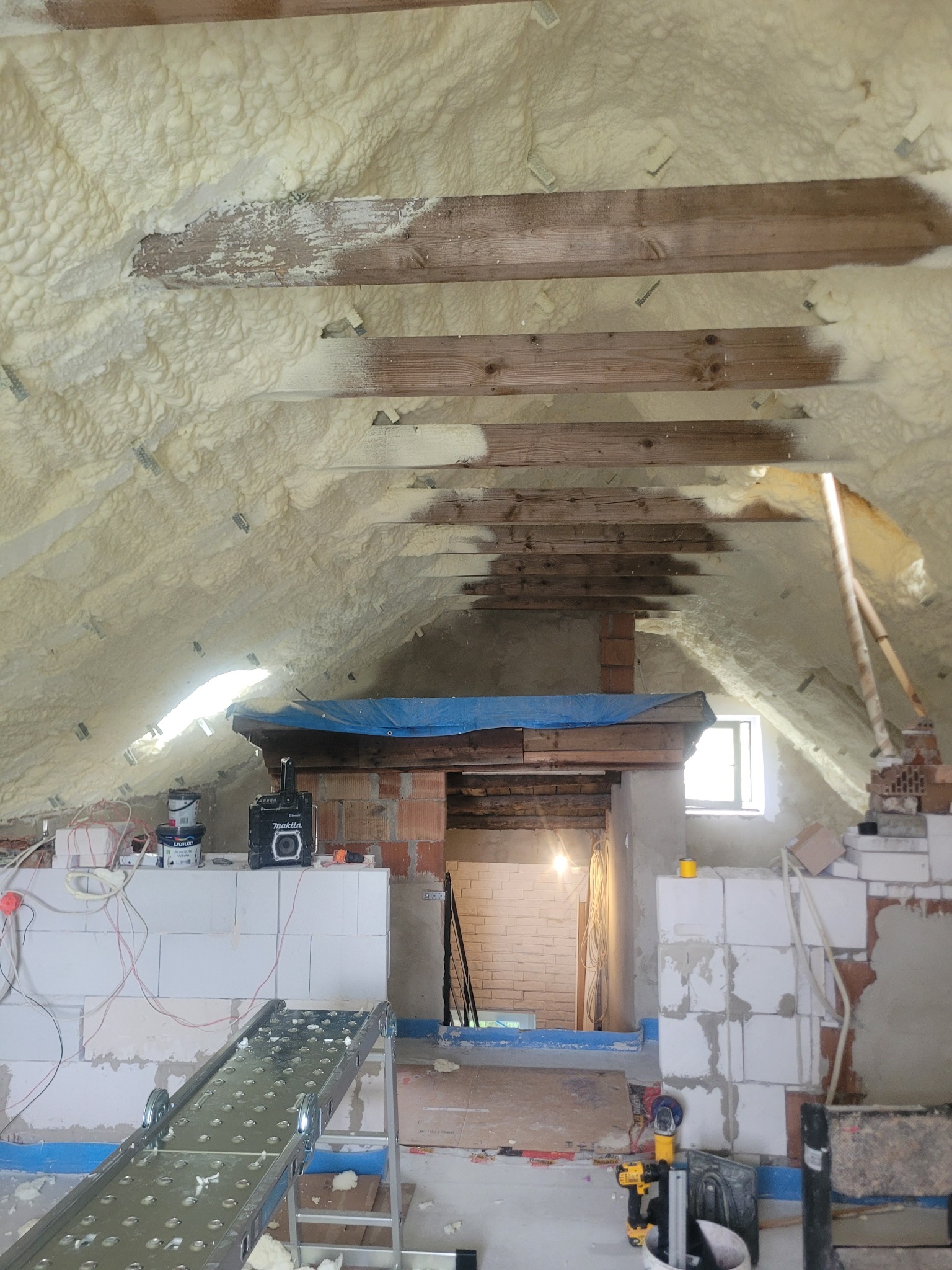 Poddasze ocieplone pianą PUR, widoczne drewniane belki konstrukcyjne. Na podłodze drabina z platformą roboczą, ściany z białych bloczków. Okno w tle.