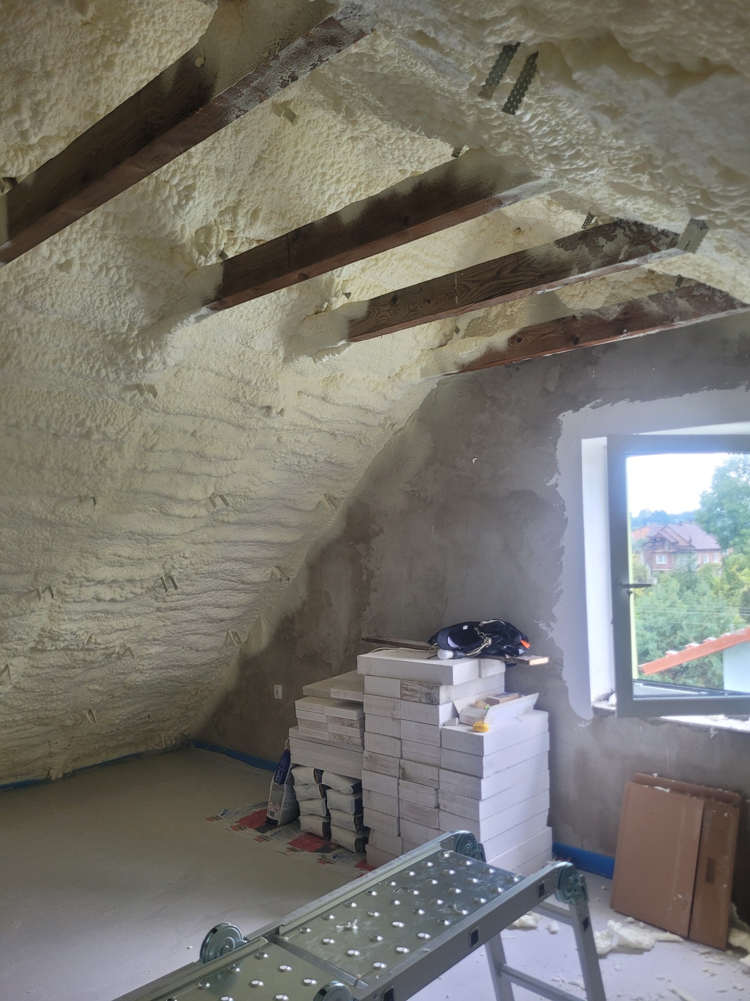 Poddasze po izolacji pianą PUR, widoczne drewniane belki i okno. Na podłodze materiały budowlane i drabina. Izolacja wykonana natryskowo, pokrywa skośne ściany.