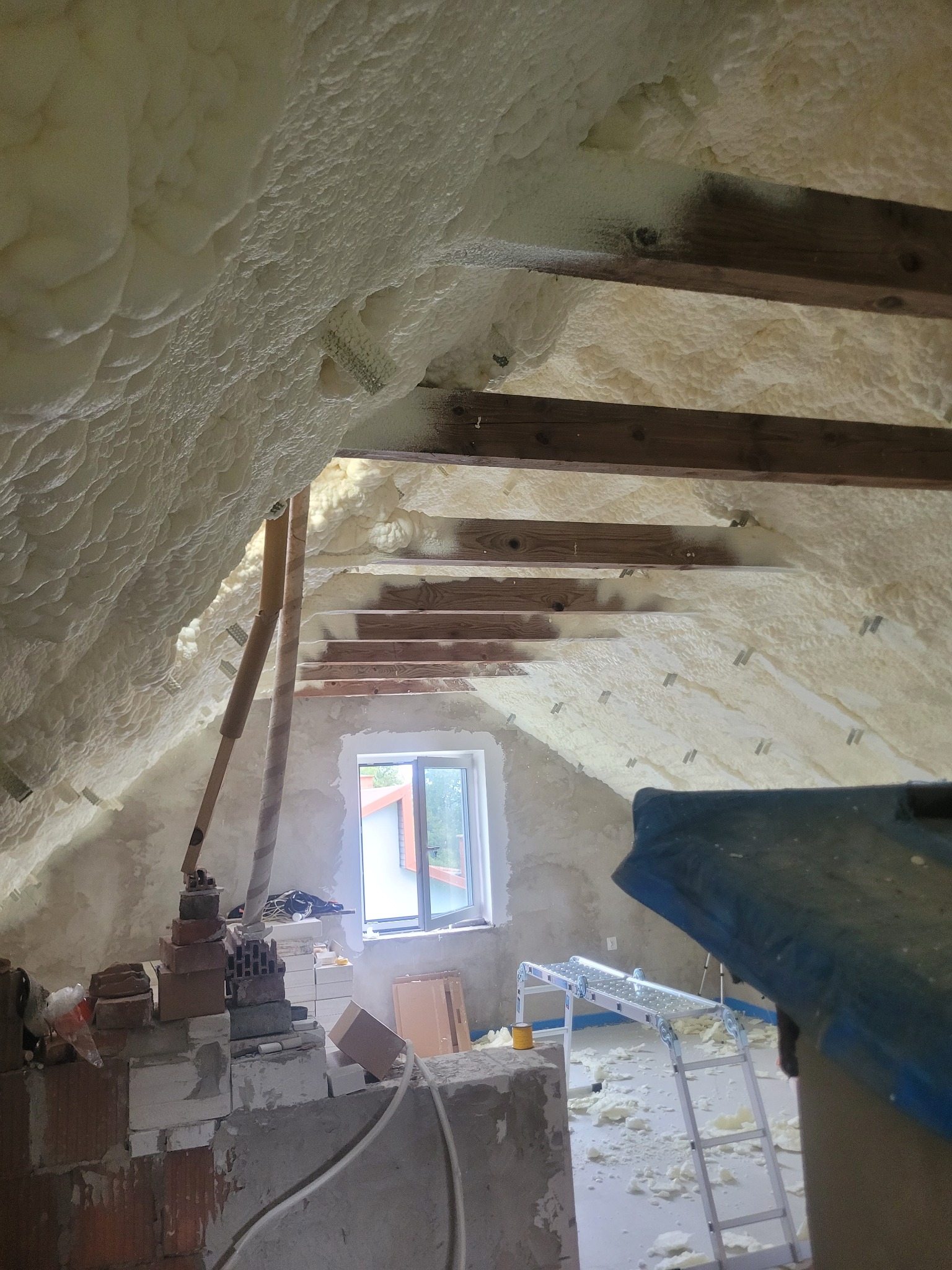 Poddasze ocieplone pianą PUR z widocznymi belkami stropowymi i oknem. Na podłodze resztki piany, cegły i drabina. Ocieplenie poddasza pianką poliuretanową.