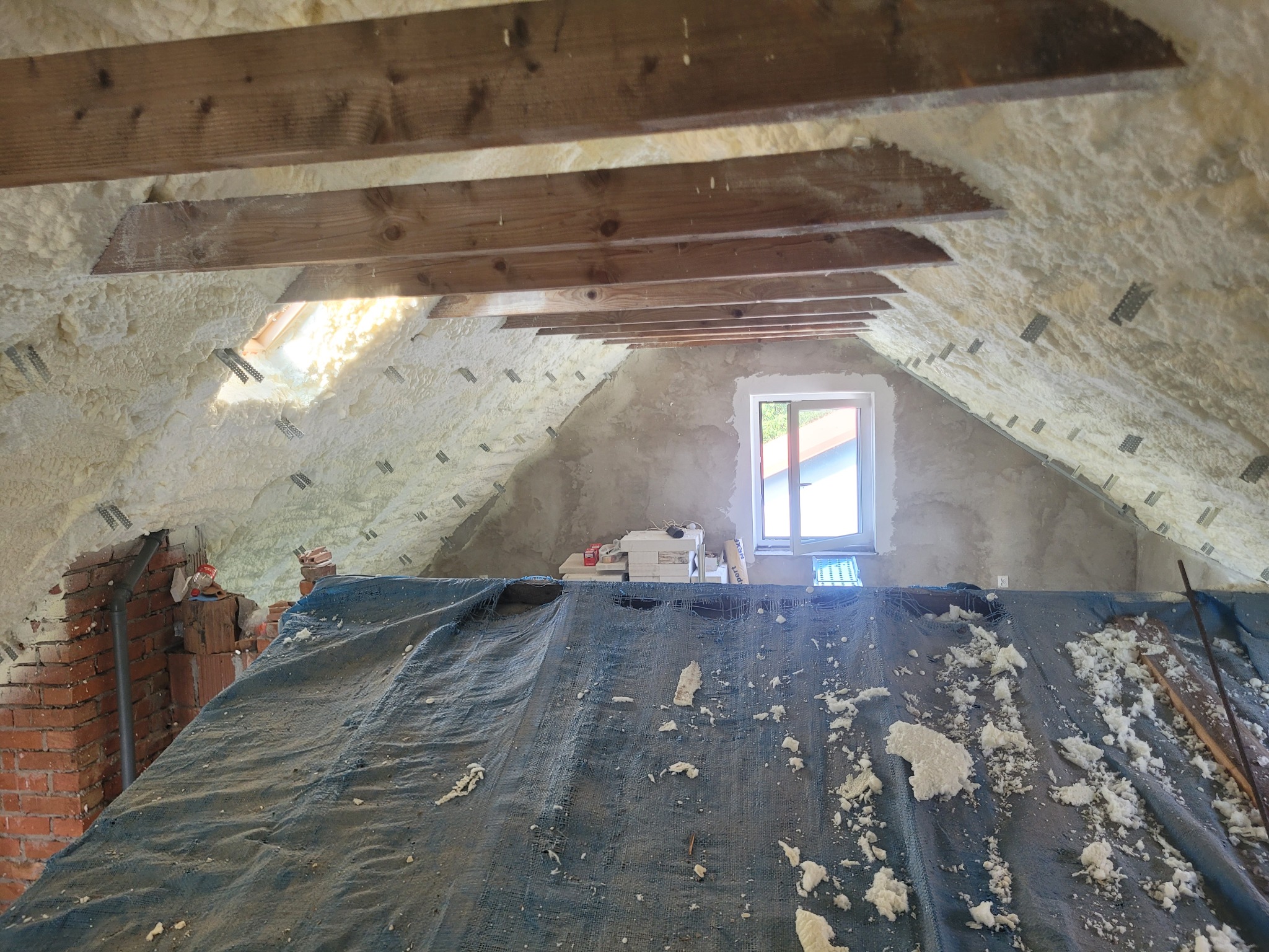 Poddasze po ociepleniu pianą PUR, widoczne drewniane elementy konstrukcyjne i okno. Część poddasza zakryta plandeką z resztkami piany. Widok z dołu.