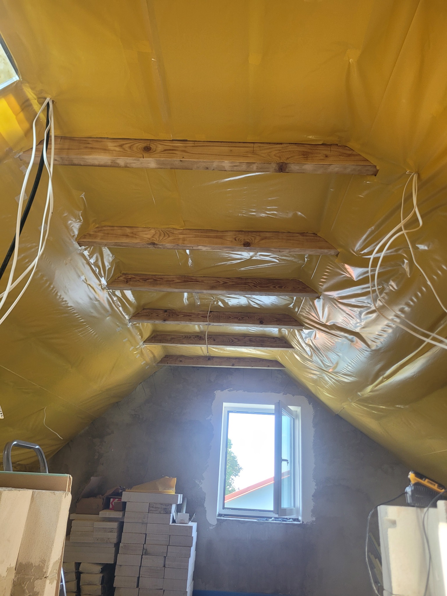 Poddasze w trakcie adaptacji: żółta folia izolacyjna, drewniane belki, okno dachowe. Widoczne materiały budowlane i narzędzia. Etap przygotowania do wykończenia.