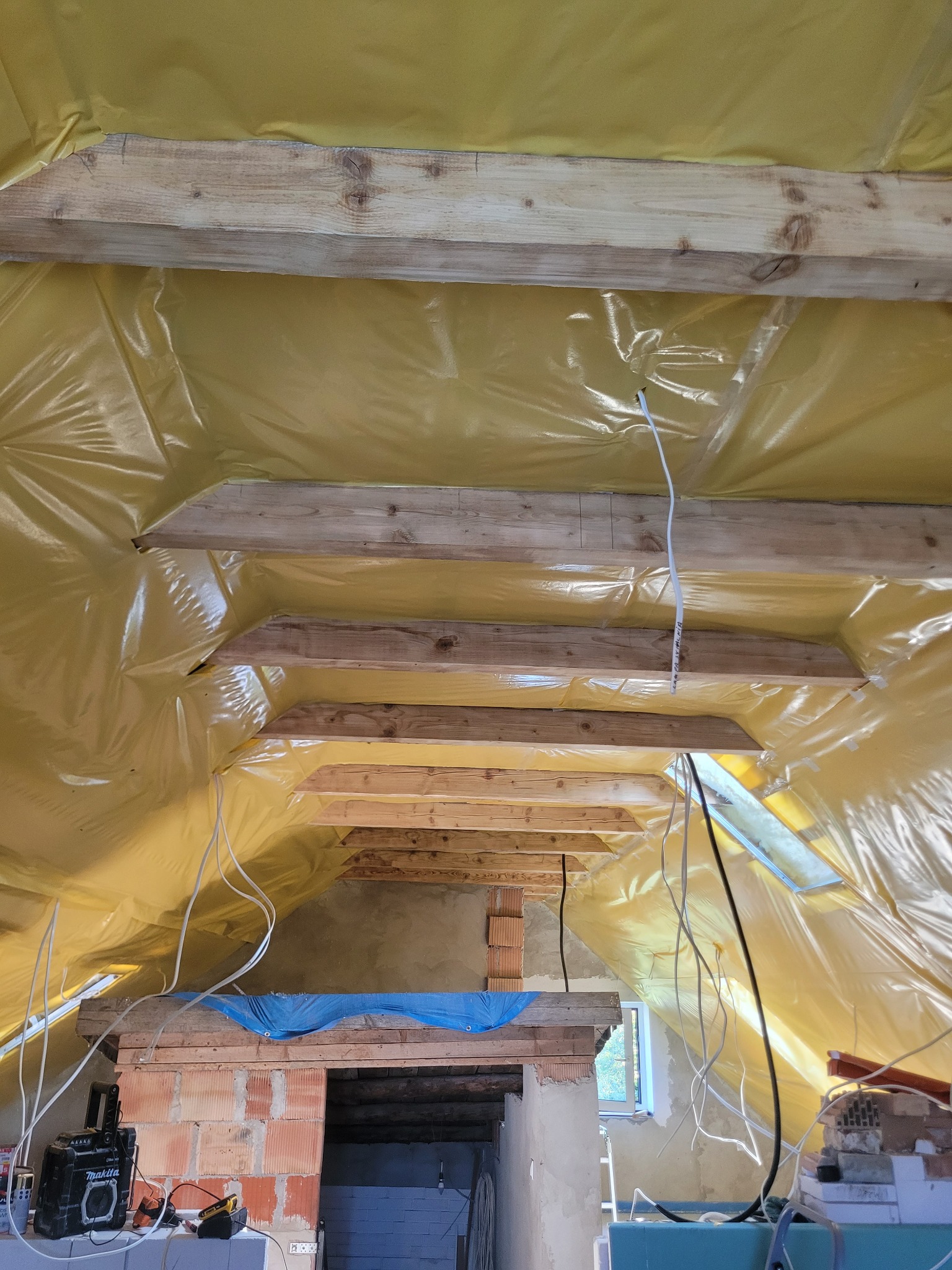 Poddasze w trakcie prac: widoczne drewniane belki stropowe, folia izolacyjna w kolorze żółtym, instalacja elektryczna i fragment murowanej konstrukcji. Prace wykończeniowe.