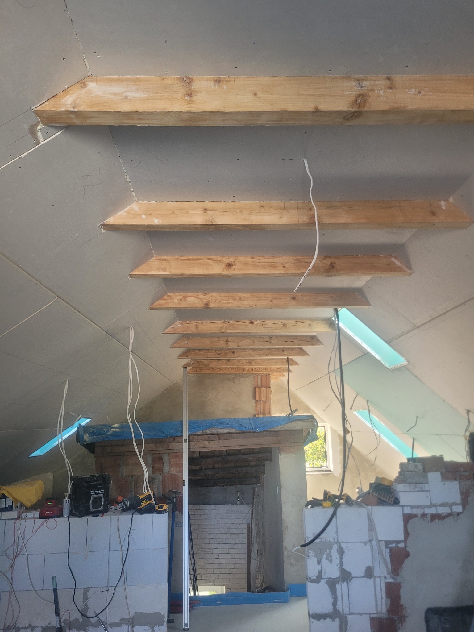 Poddasze w trakcie prac wykończeniowych: płyty gipsowo-kartonowe, drewniane belki, widoczne okno dachowe i instalacje elektryczne. Fragment muru z cegieł.
