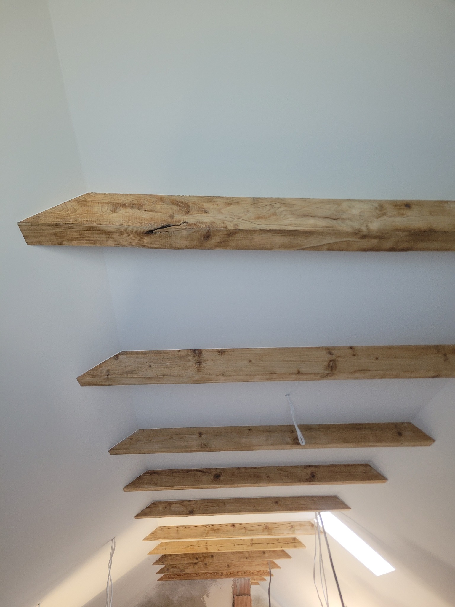 Wykończone drewniane belki stropowe na poddaszu, widok z dołu. Element dekoracyjny w jasnym wnętrzu. Widoczne kable i fragment ściany z cegły.