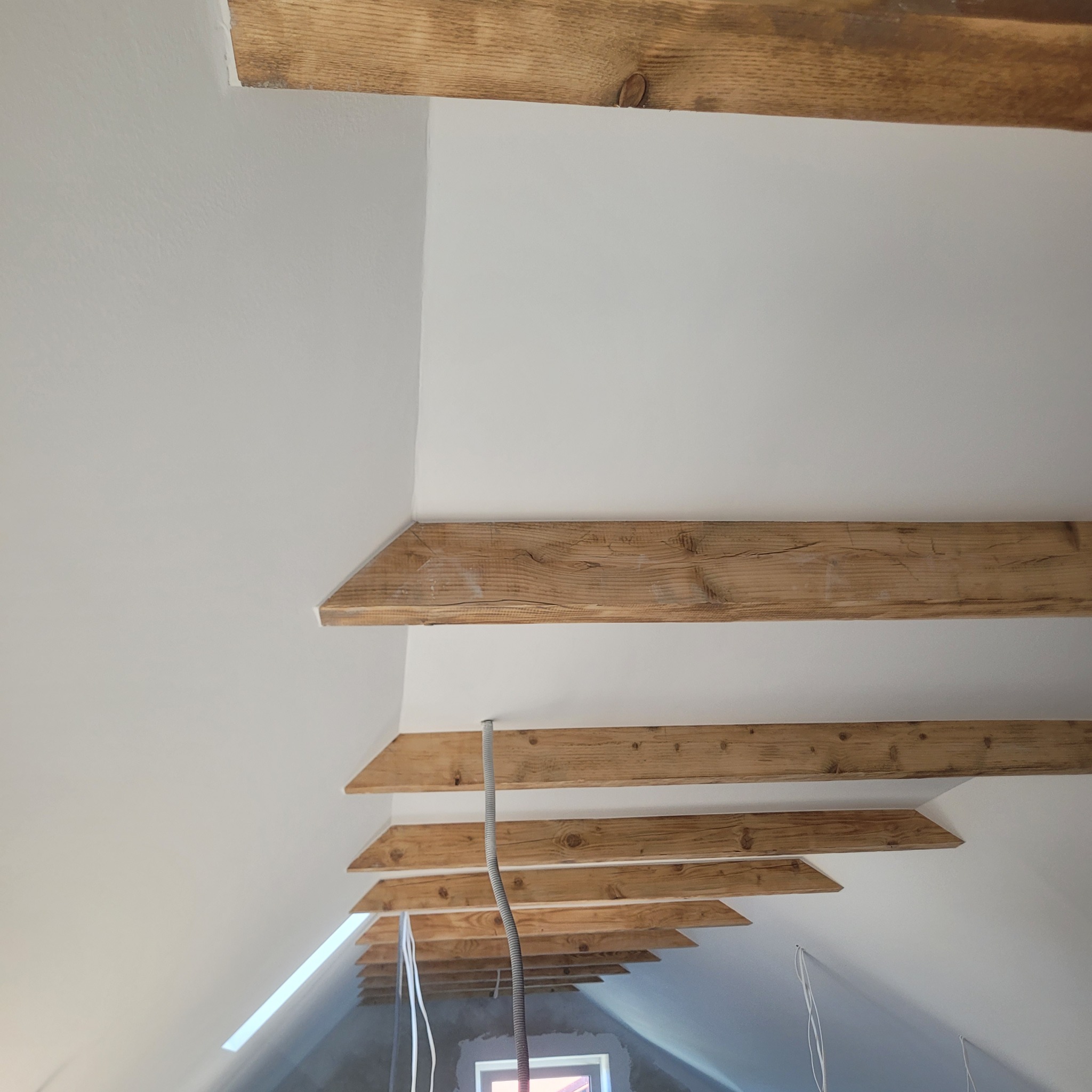 Białe poddasze z drewnianymi belkami stropowymi. Widoczne kable i rury instalacyjne. Ujęcie z dołu, podkreślające strukturę i detale wykończenia.