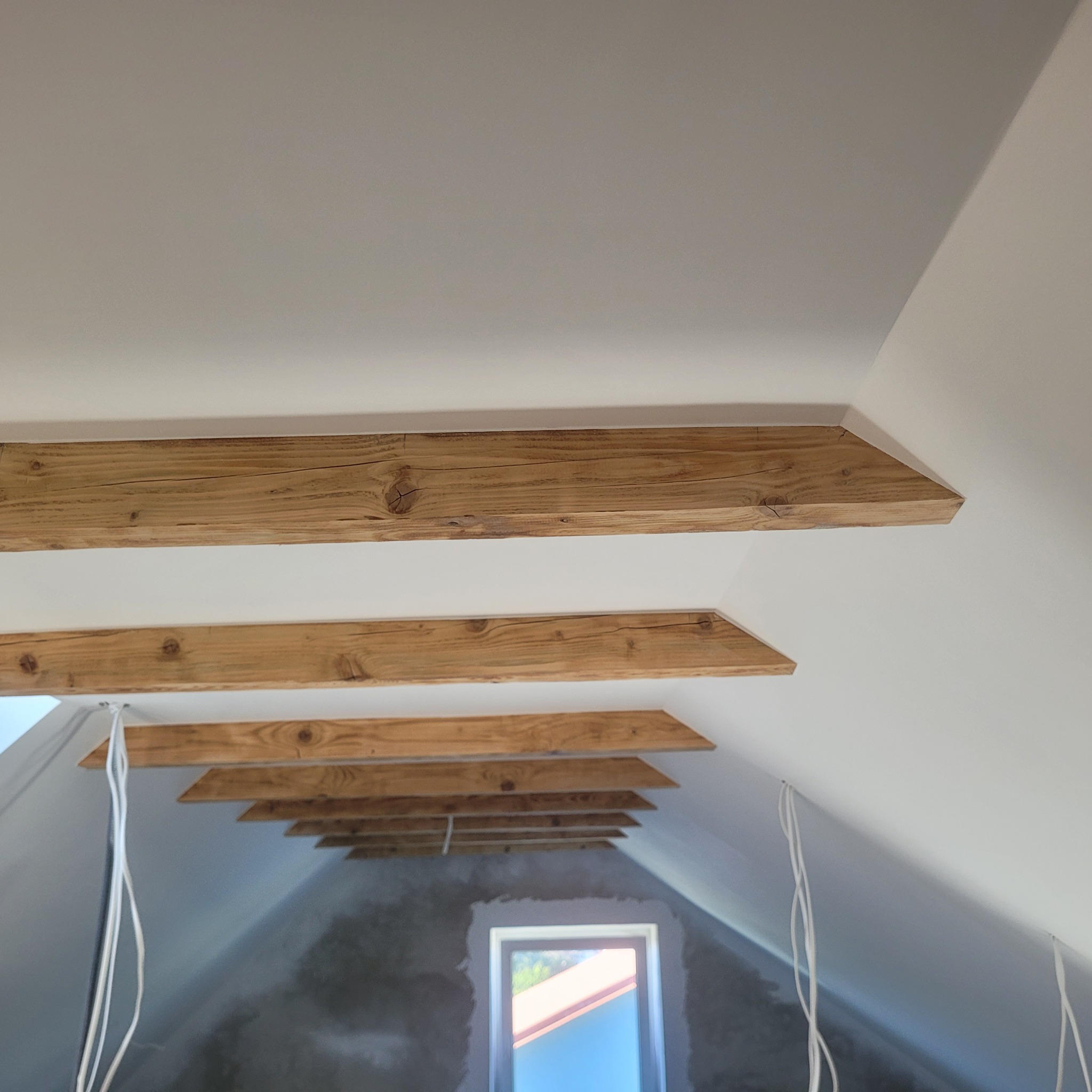 Wykończone drewniane belki stropowe w ocieplonym poddaszu w Kamionnej. Widoczne okno i przewody elektryczne.