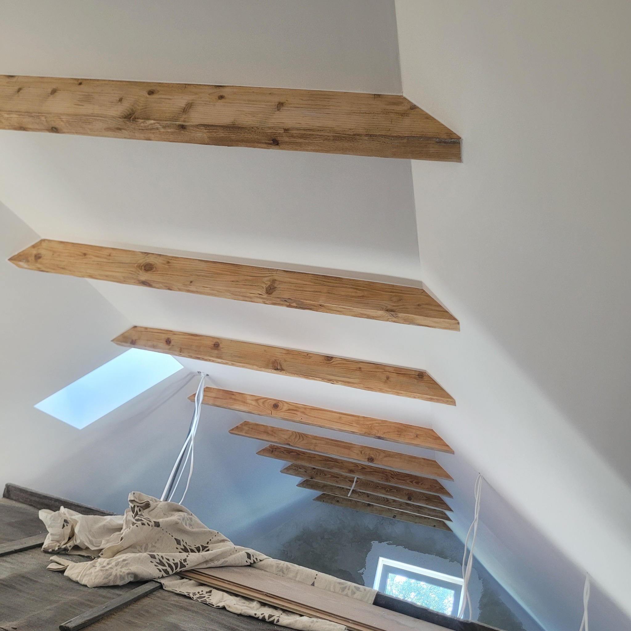 Poddasze z drewnianymi belkami stropowymi i oknem dachowym. Widoczne kable i materiały budowlane. Świeżo pomalowane ściany w jasnym odcieniu.