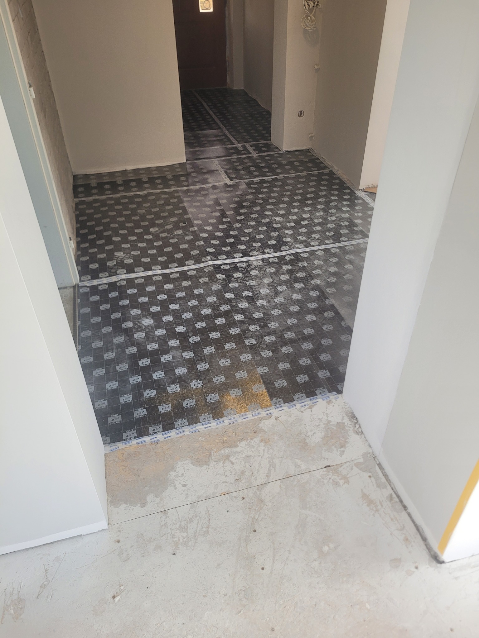 Przygotowanie podłoża pod panele: rozłożona mata wygłuszająca z nadrukiem w korytarzu, widok z progu. Białe ściany, surowa podłoga w tle.