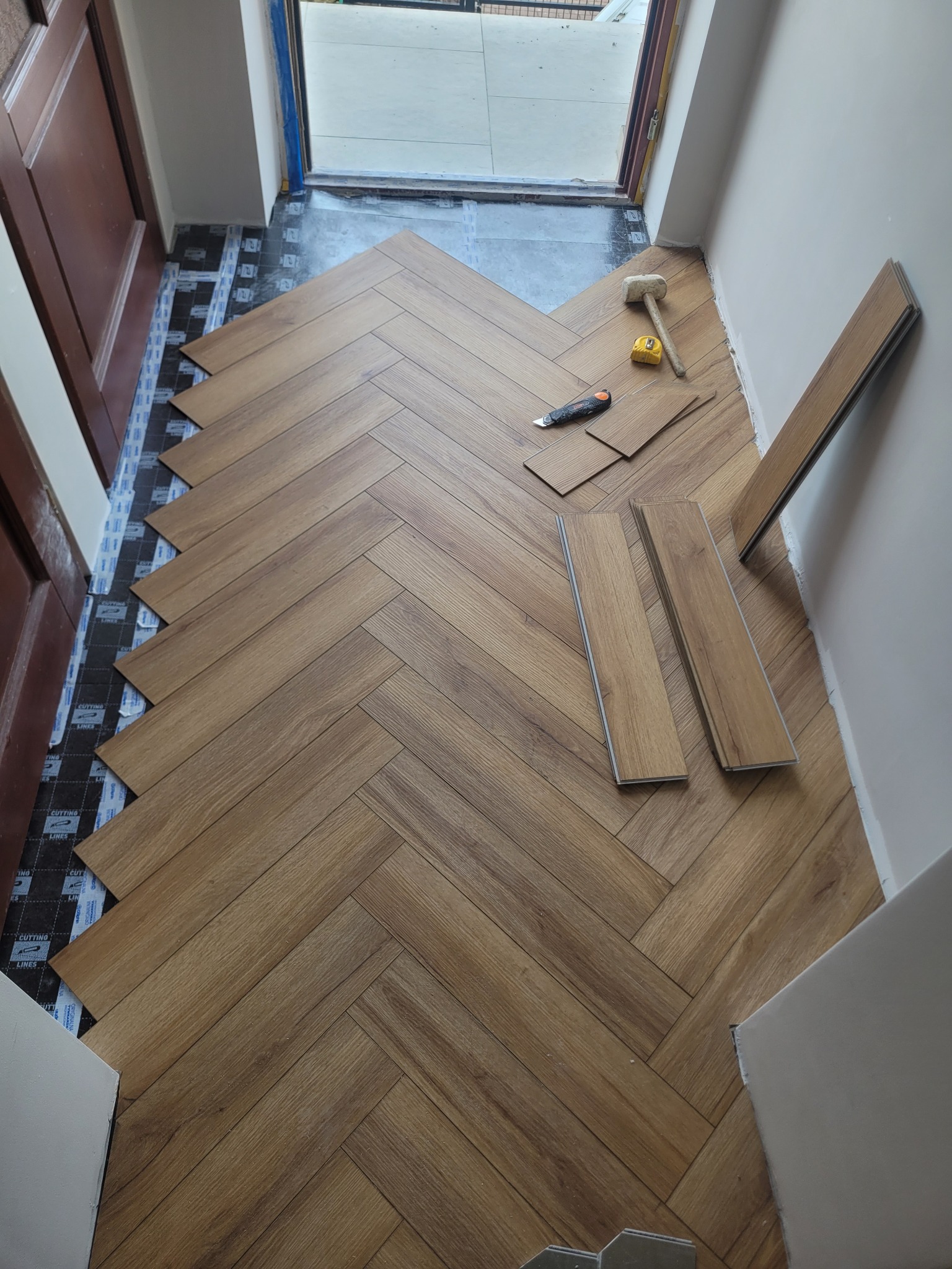 Układanie paneli podłogowych w jodełkę w przedpokoju. Widoczne narzędzia: gumowy młotek, miarka, nóż. Częściowo ułożona podłoga na podkładzie izolacyjnym.