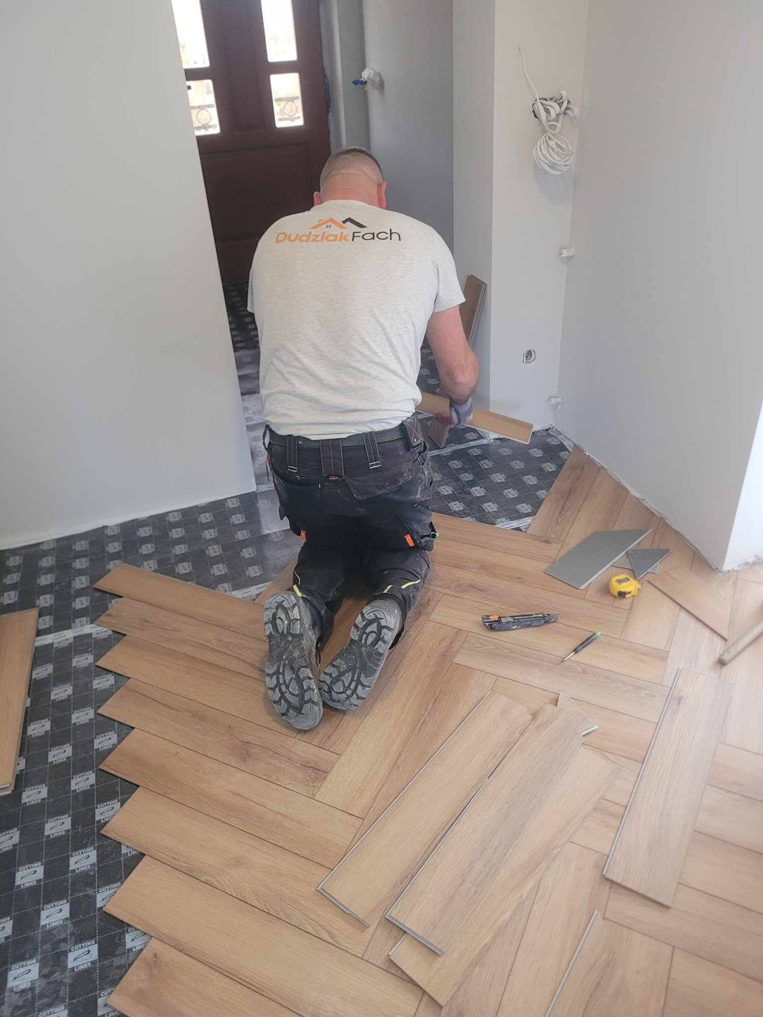 Układanie paneli podłogowych w jodełkę. Widoczny pracownik na kolanach, narzędzia: nóż, miarka. Częściowo ułożona podłoga na podkładzie. Jasne wnętrze.