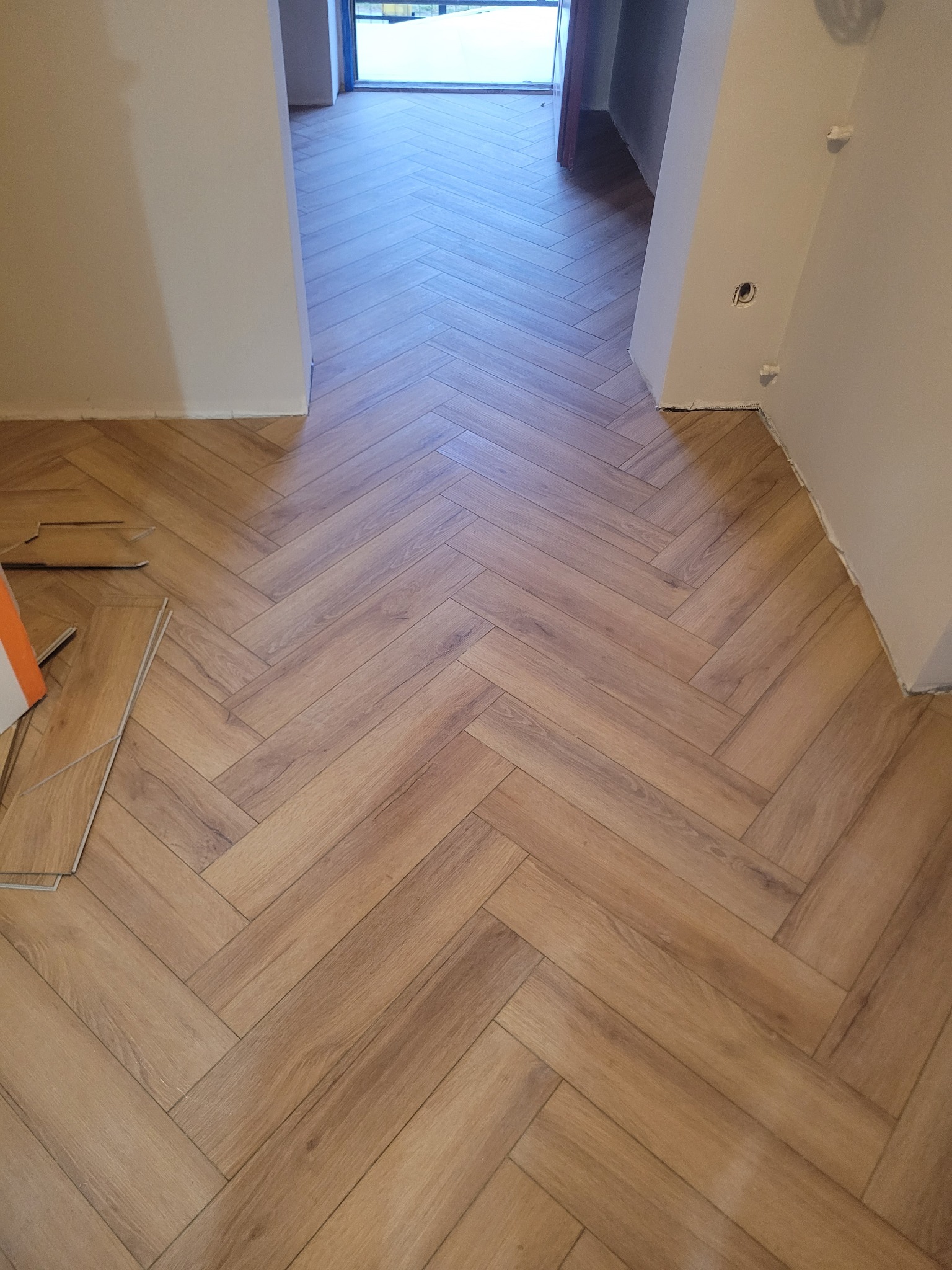 Ukończona podłoga z paneli ułożonych w jodełkę, widoczne niedocięte panele przy ścianie. Jasne ściany i widok na dalsze pomieszczenie.