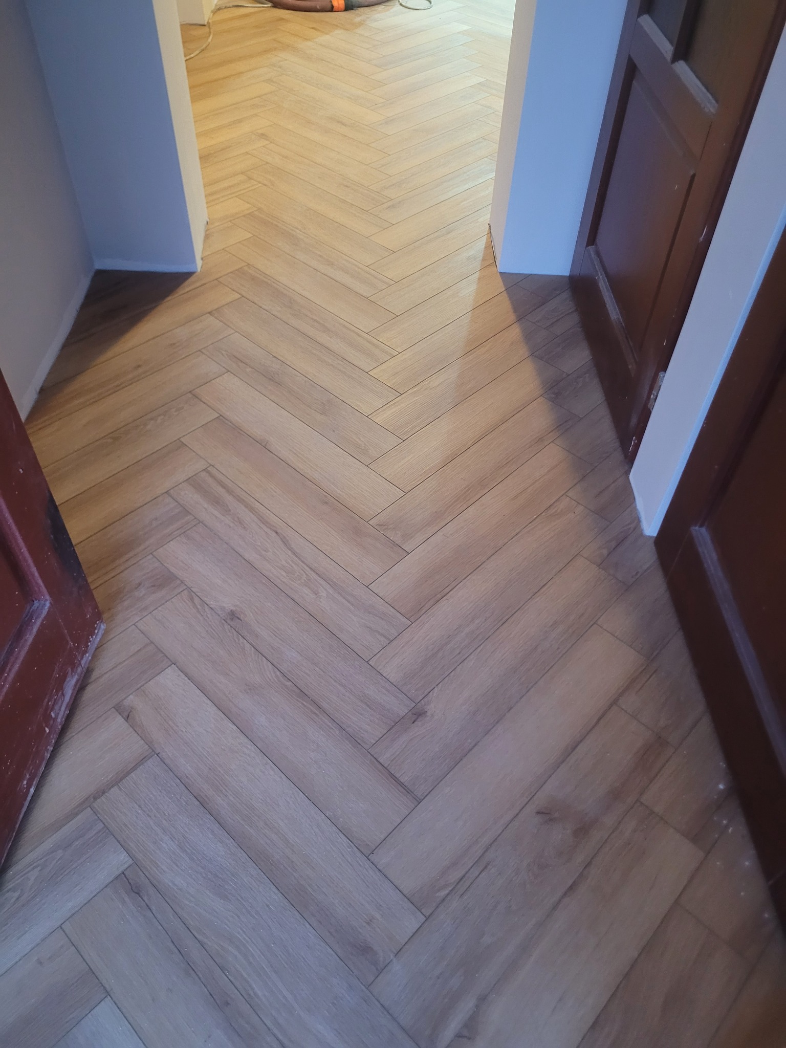 Ułożona podłoga z paneli w jodełkę, widok z góry na korytarz z otwartymi drzwiami po bokach. Jasne panele imitują drewno, tworząc elegancki wzór.