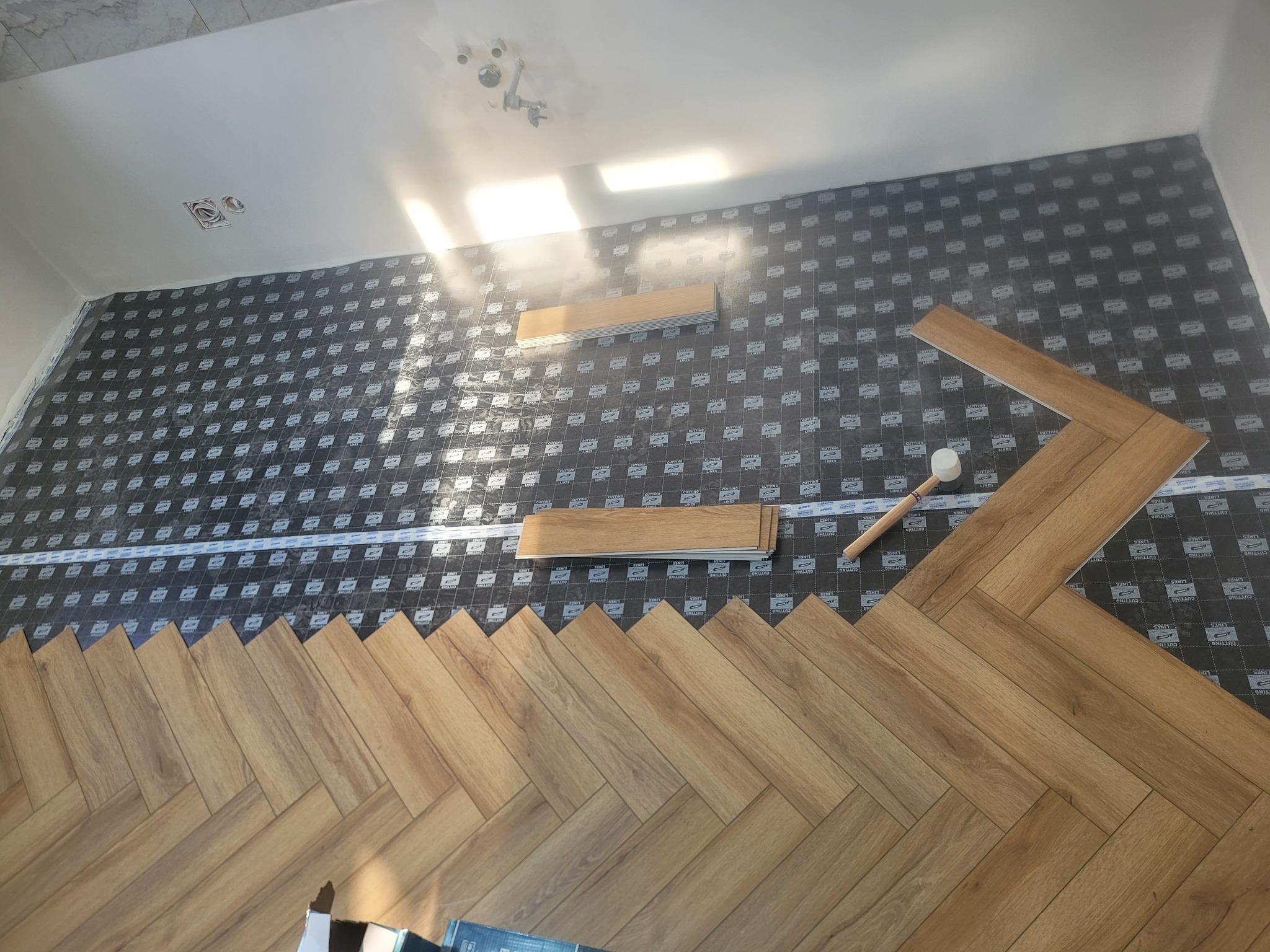 Układanie paneli podłogowych w jodełkę na podkładzie izolacyjnym. Widoczny gumowy młotek i paczka paneli. Ujęcie z góry, w trakcie montażu.