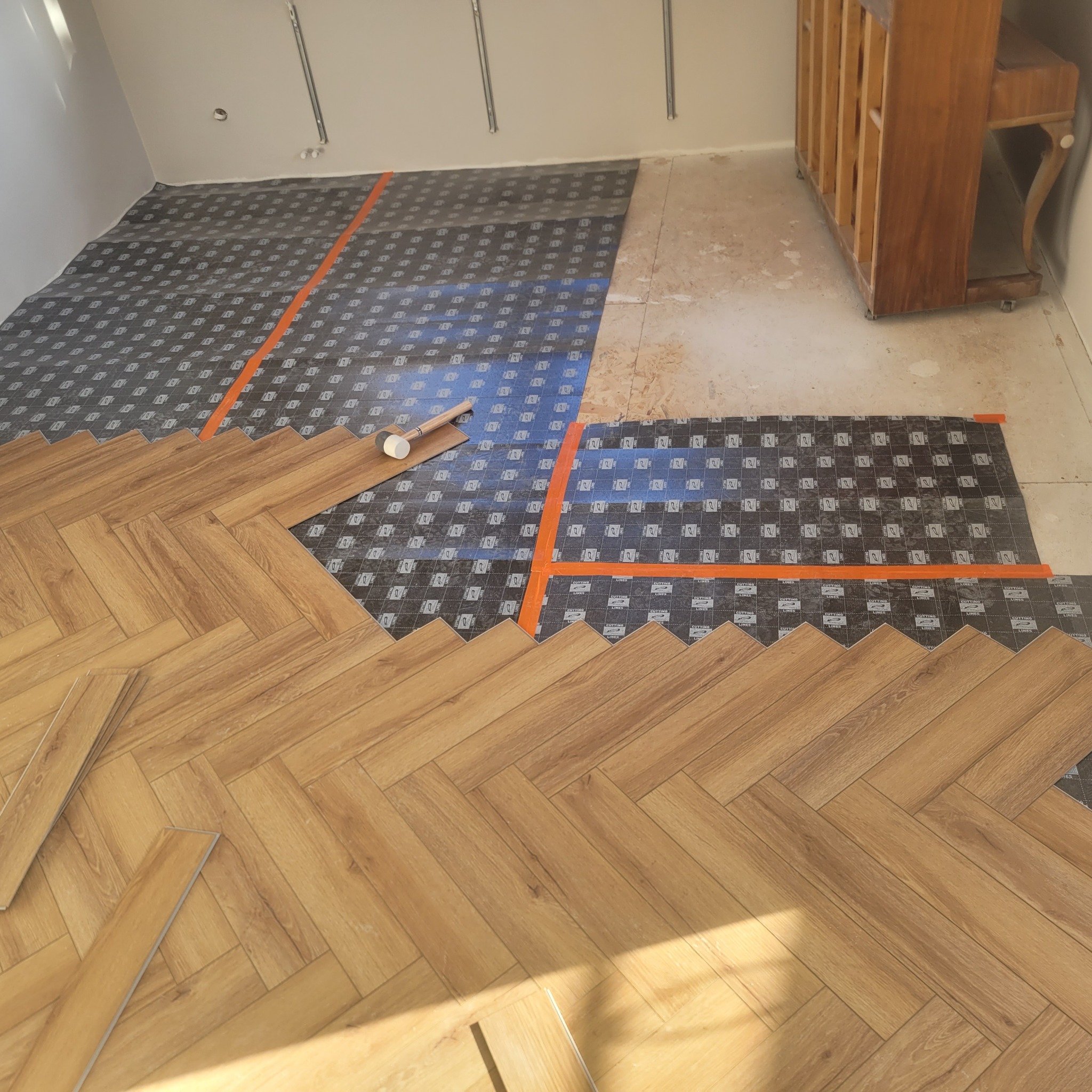 Układanie paneli podłogowych w jodełkę na podkładzie z folii. Częściowo ułożona podłoga z widocznym gumowym młotkiem. W tle fragment szafki.