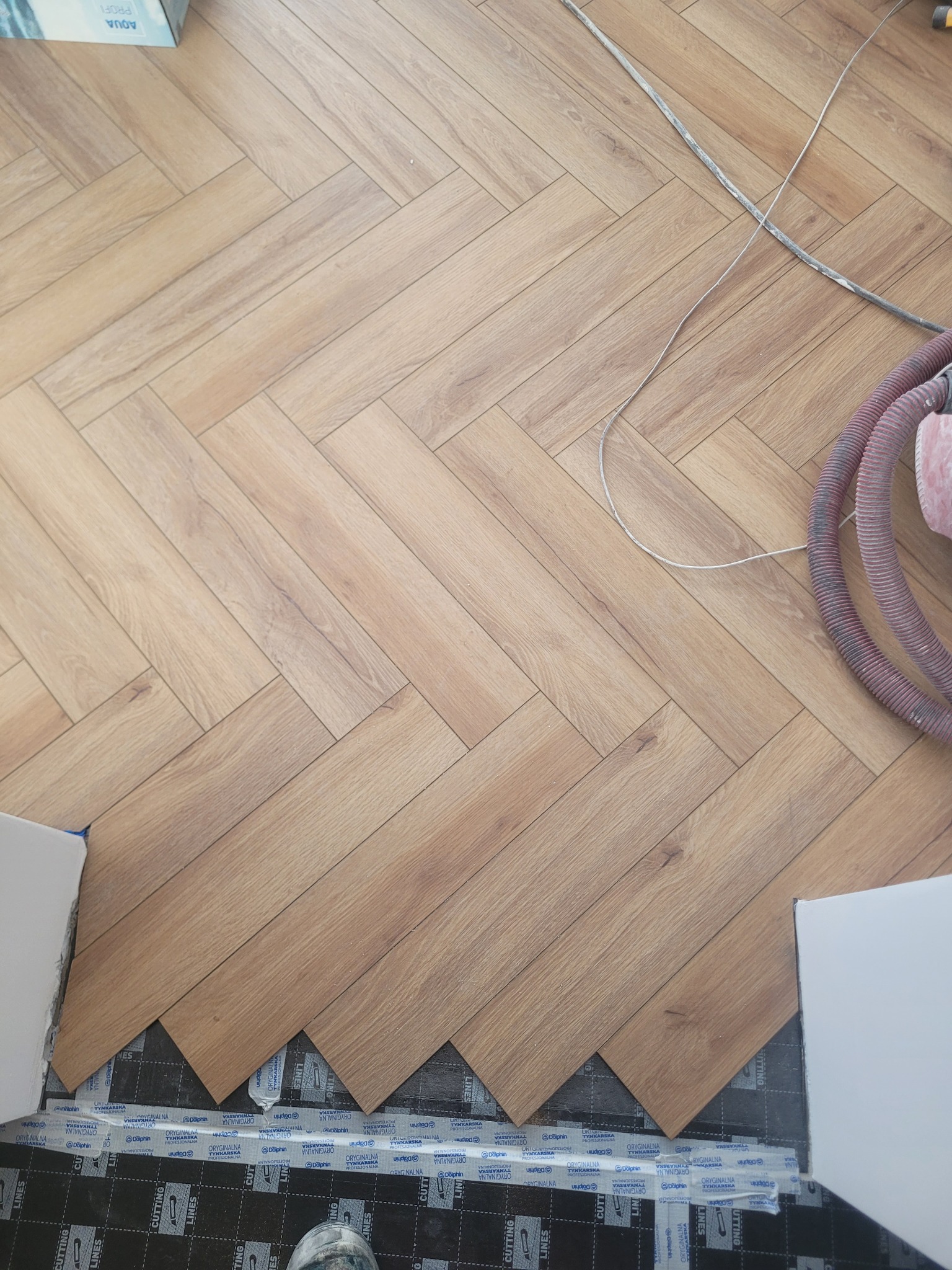 Układanie paneli podłogowych w jodełkę. Widoczne panele w odcieniach brązu, podkład pod panele z nadrukiem, fragment odkurzacza przemysłowego i listwa przypodłogowa.