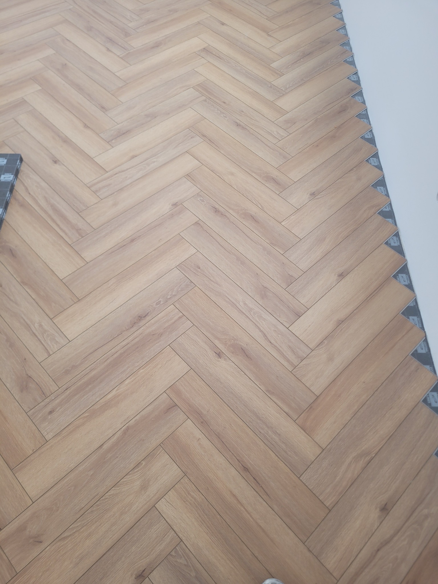 Ułożone panele podłogowe w jodełkę, widoczne przy ścianie podkładki dystansowe. Jasne, drewnopodobne panele tworzą wzór na podłodze.