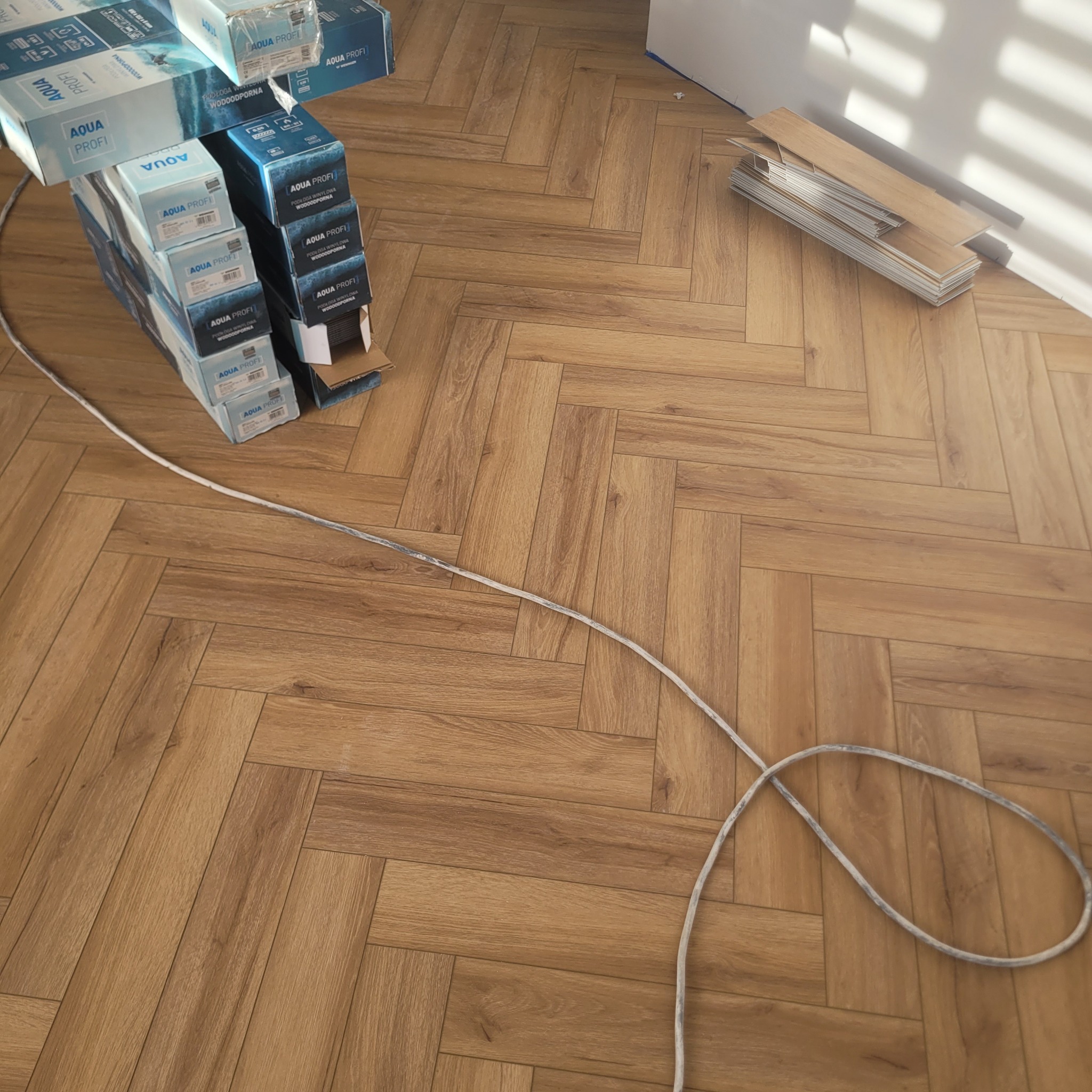 Ułożona podłoga winylowa w jodełkę, z paczkami paneli Aqua Profi i listwami przypodłogowymi przy ścianie. Widoczne światło słoneczne.