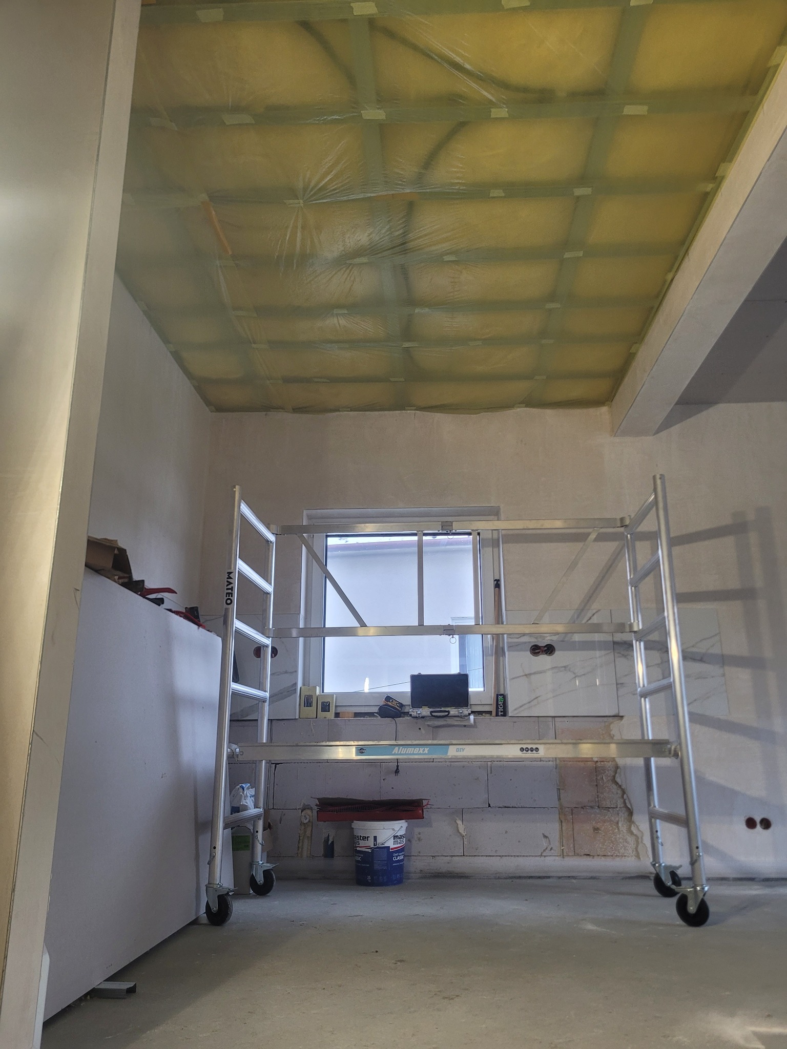 Sufit z izolacją termiczną zabezpieczoną folią, widoczny z poziomu rusztowania w trakcie prac wykończeniowych. W tle okno i ściana z widocznymi śladami prac.