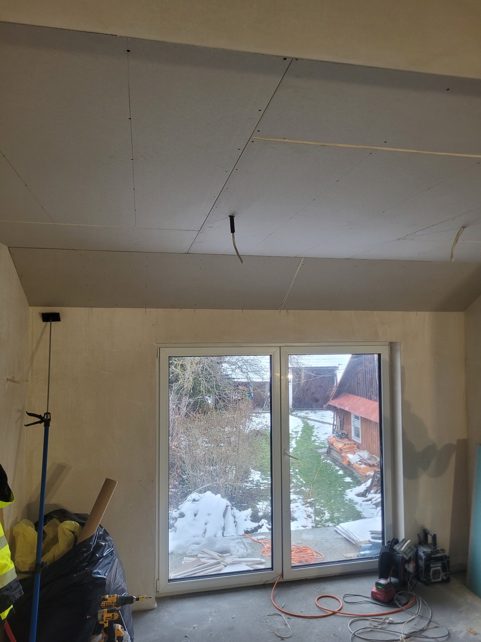 Płyty karton-gips na suficie w trakcie montażu, widoczne narzędzia i okno z widokiem na zasypany śniegiem ogród. Prace wykończeniowe w pomieszczeniu.