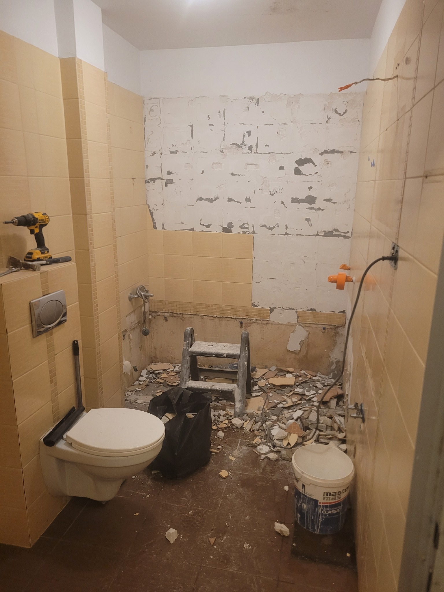 Demontaż płytek w łazience: widoczne ślady po usuniętych płytkach, drabina, narzędzia i gruz. Częściowo zachowane beżowe płytki na ścianach.