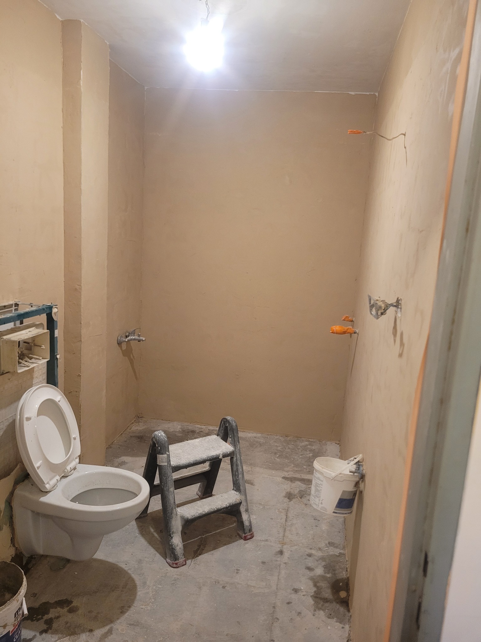 Łazienka w trakcie prac wykończeniowych: ściany po gładzi, zamontowana toaleta, drabina malarska, widoczne instalacje. Oświetlenie sufitowe.
