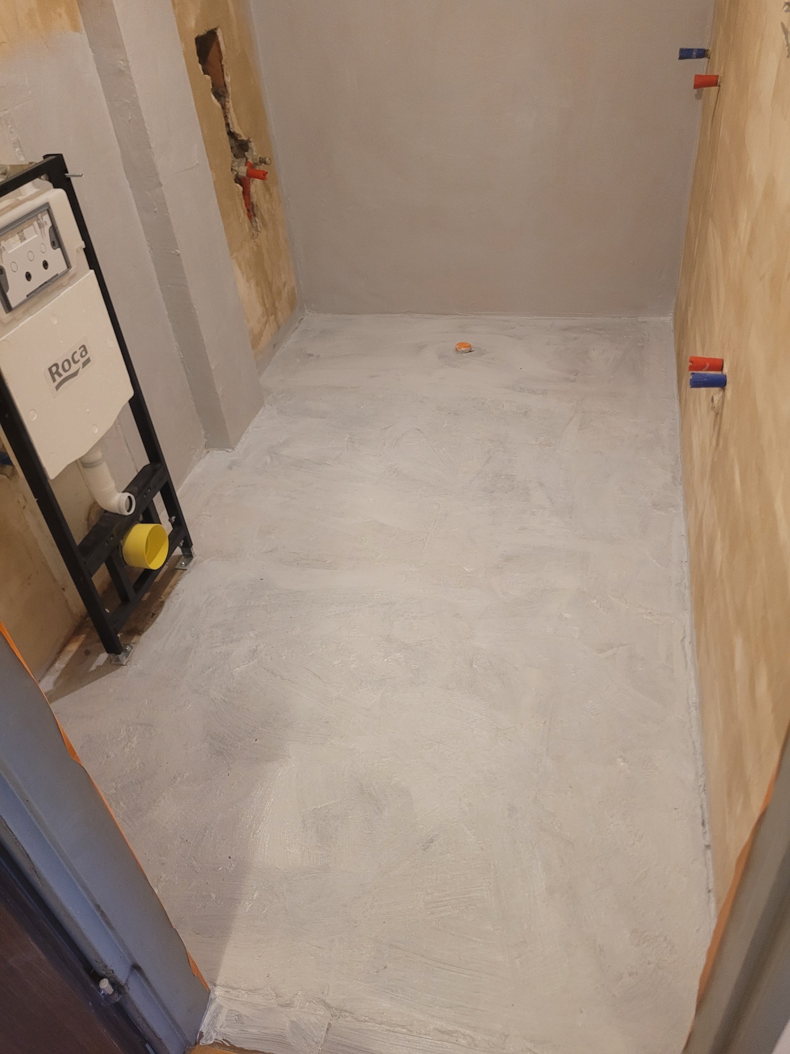 Przygotowana do dalszych prac łazienka: zagruntowane ściany, wylewka na podłodze, zamontowany stelaż podtynkowy WC Roca z żółtym elementem. Widoczne wystające rury.