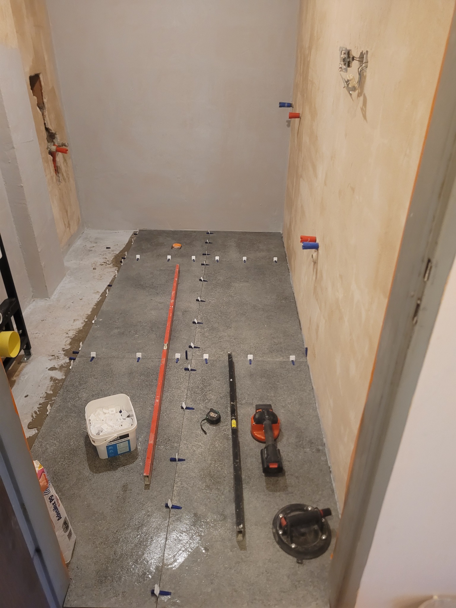 Szare płytki podłogowe w trakcie układania w łazience, widoczne poziomica, przyssawka do płytek i wiadro z klinami dystansowymi. Ściany przygotowane pod wykończenie.