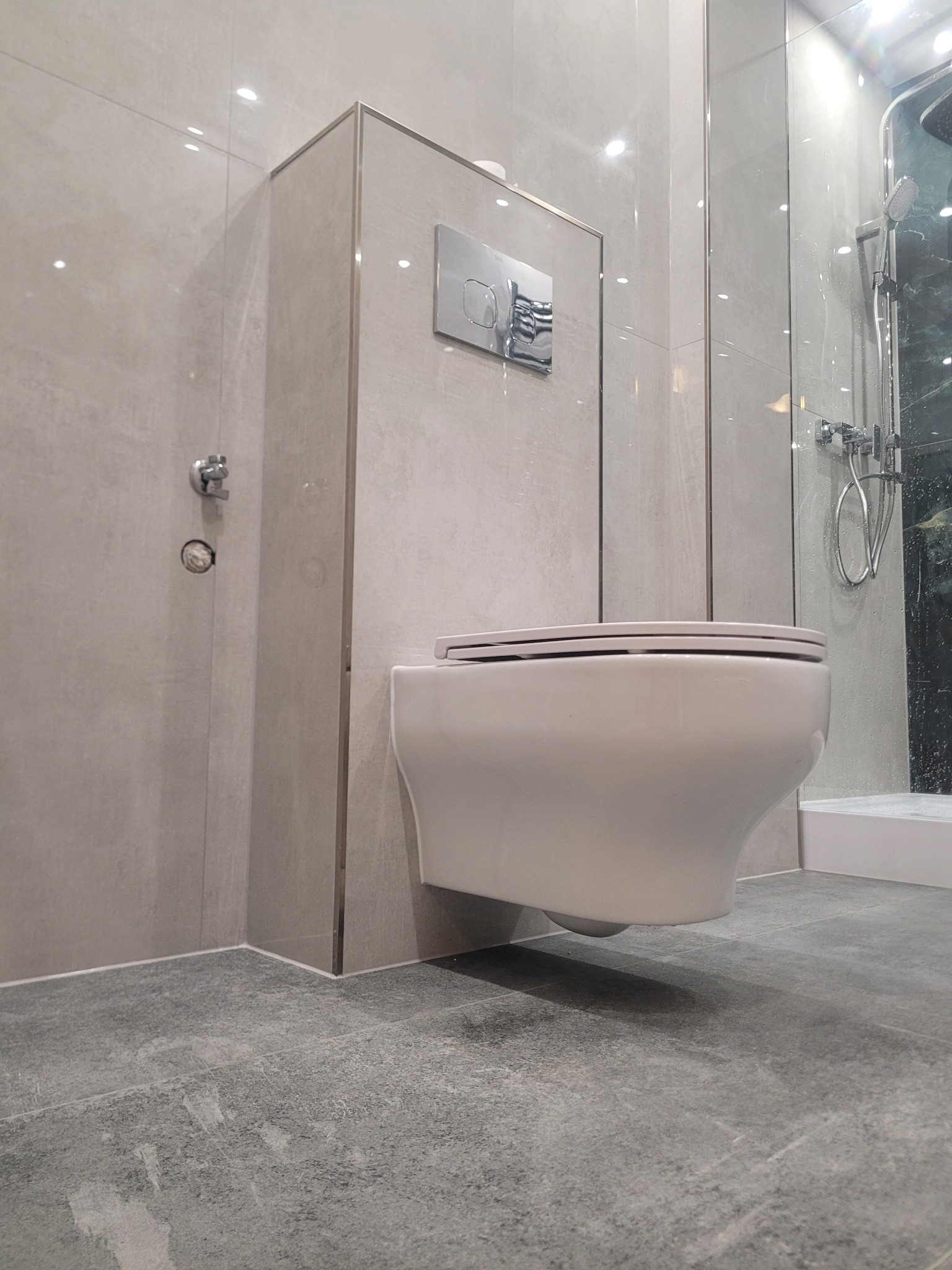 Nowoczesna łazienka z podwieszaną miską WC, szare płytki na ścianach i podłodze, chromowane detale, kabina prysznicowa ze szklaną ścianką. Minimalistyczny design.