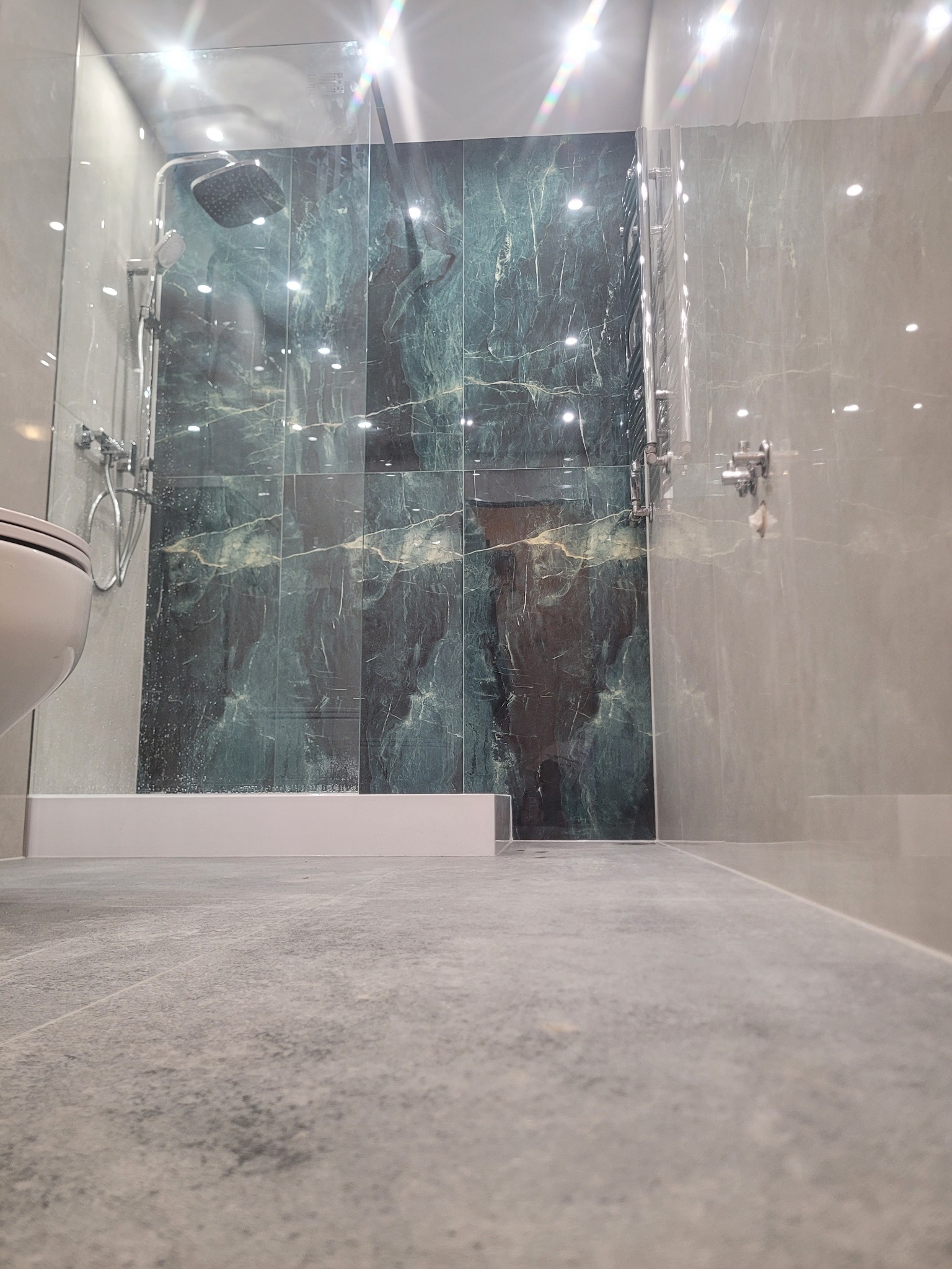 Wykończona łazienka z kabiną prysznicową i płytkami imitującymi marmur w odcieniach zieleni i szarości. Widok z poziomu podłogi, z fragmentem toalety po lewej.