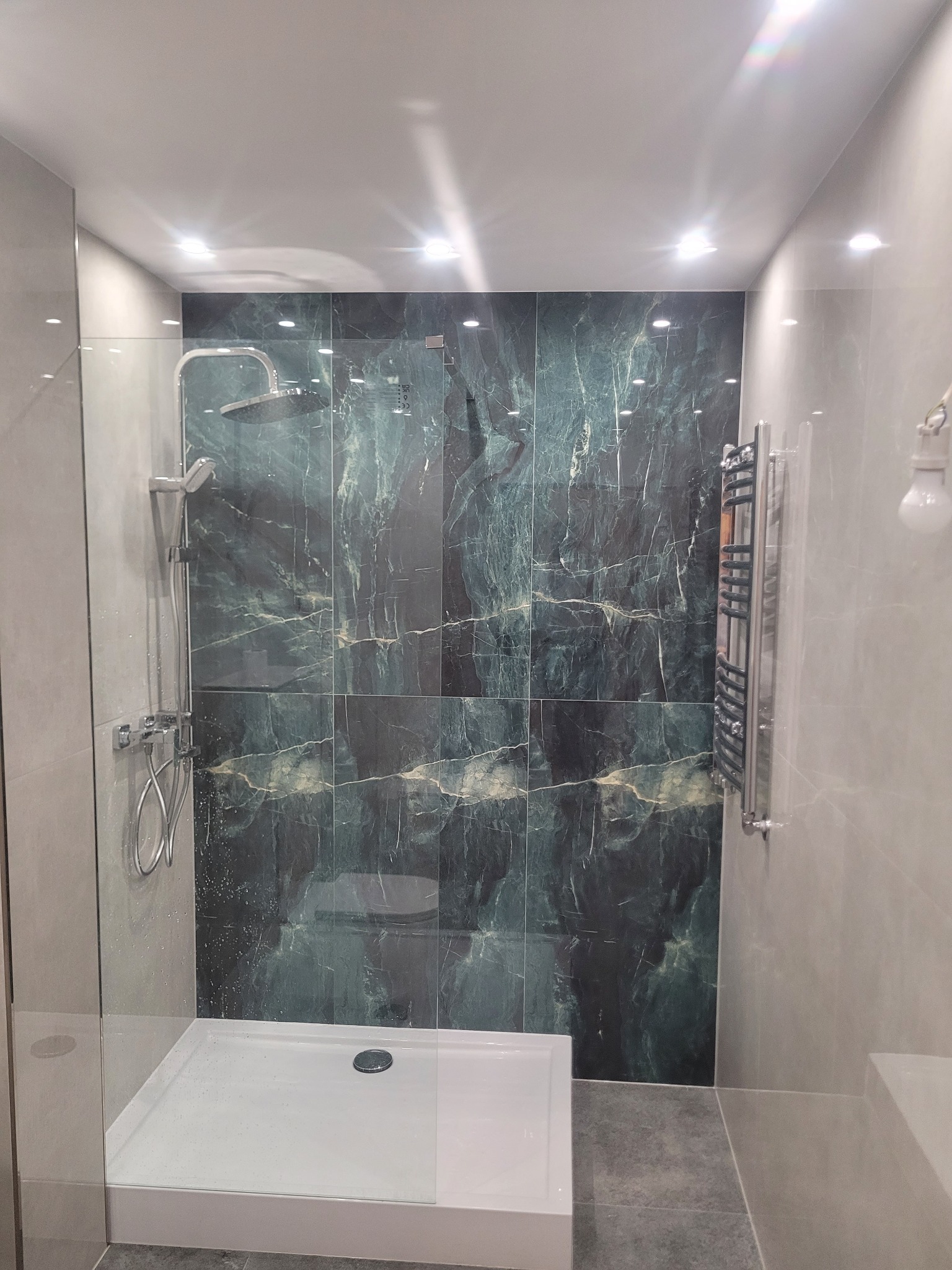Nowoczesna kabina prysznicowa z deszczownicą i płytkami imitującymi marmur w odcieniach zieleni i szarości. Widoczny grzejnik drabinkowy i oświetlenie punktowe.