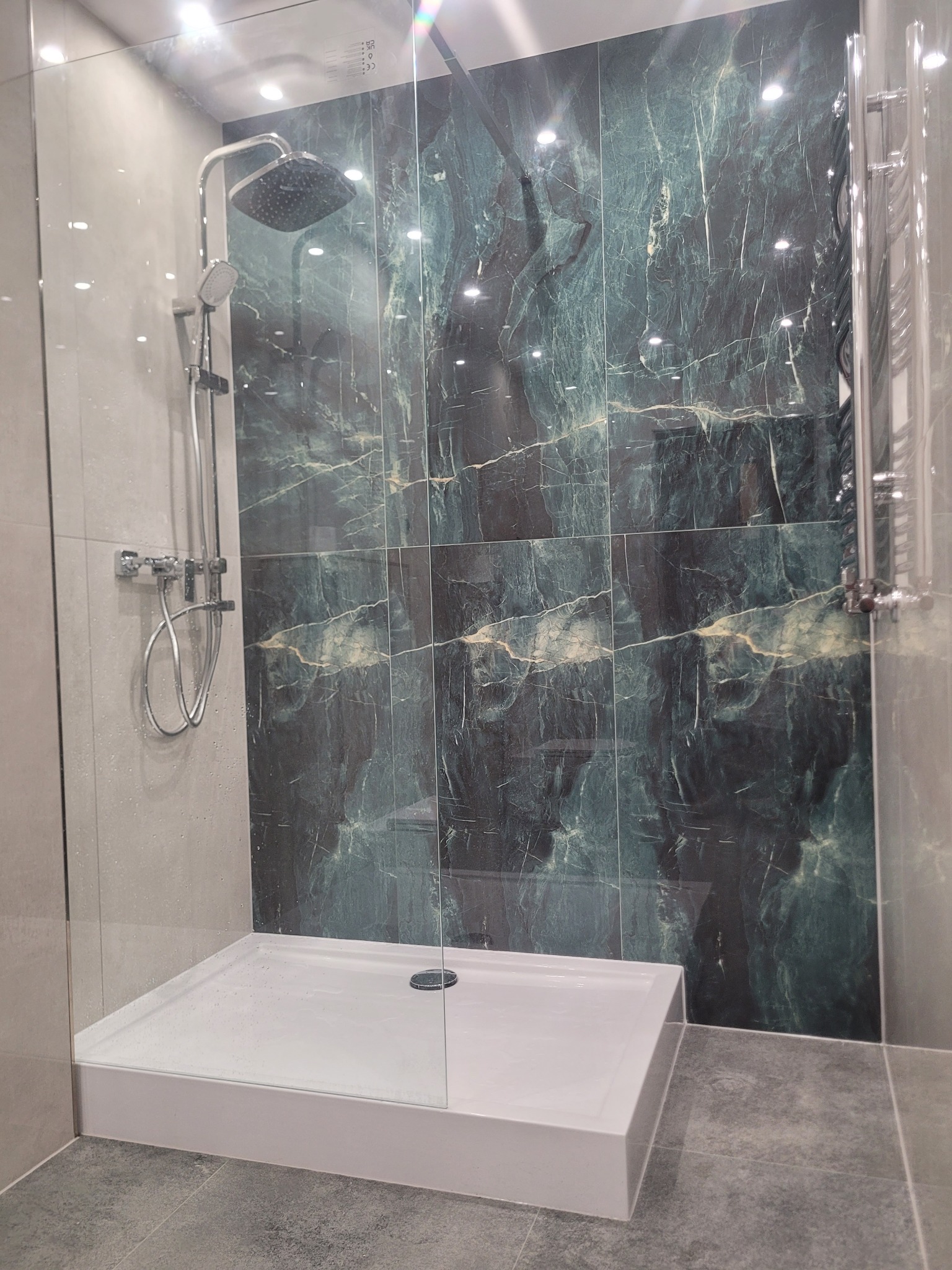 Nowoczesna kabina prysznicowa z deszczownicą i płytkami imitującymi marmur w odcieniach zieleni i szarości. Szklana ściana kabiny, biały brodzik i grzejnik drabinkowy.