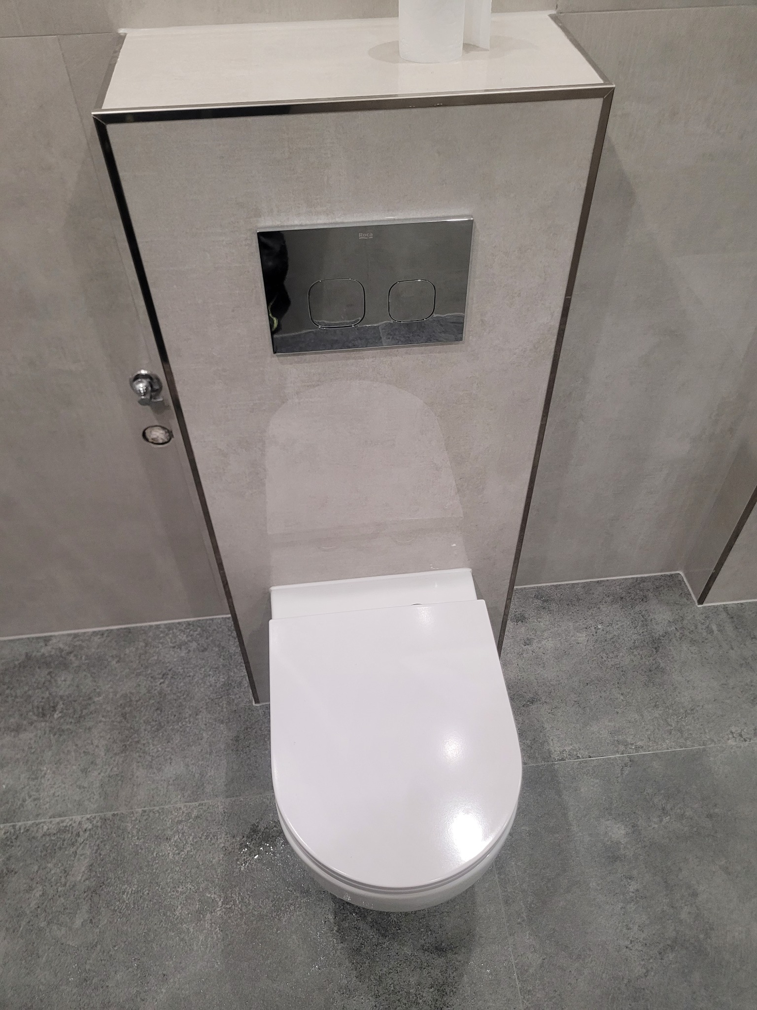 Nowoczesna toaleta podwieszana z chromowanym przyciskiem spłukującym, obudowana płytkami w odcieniach szarości. Minimalistyczny design, widok z góry.