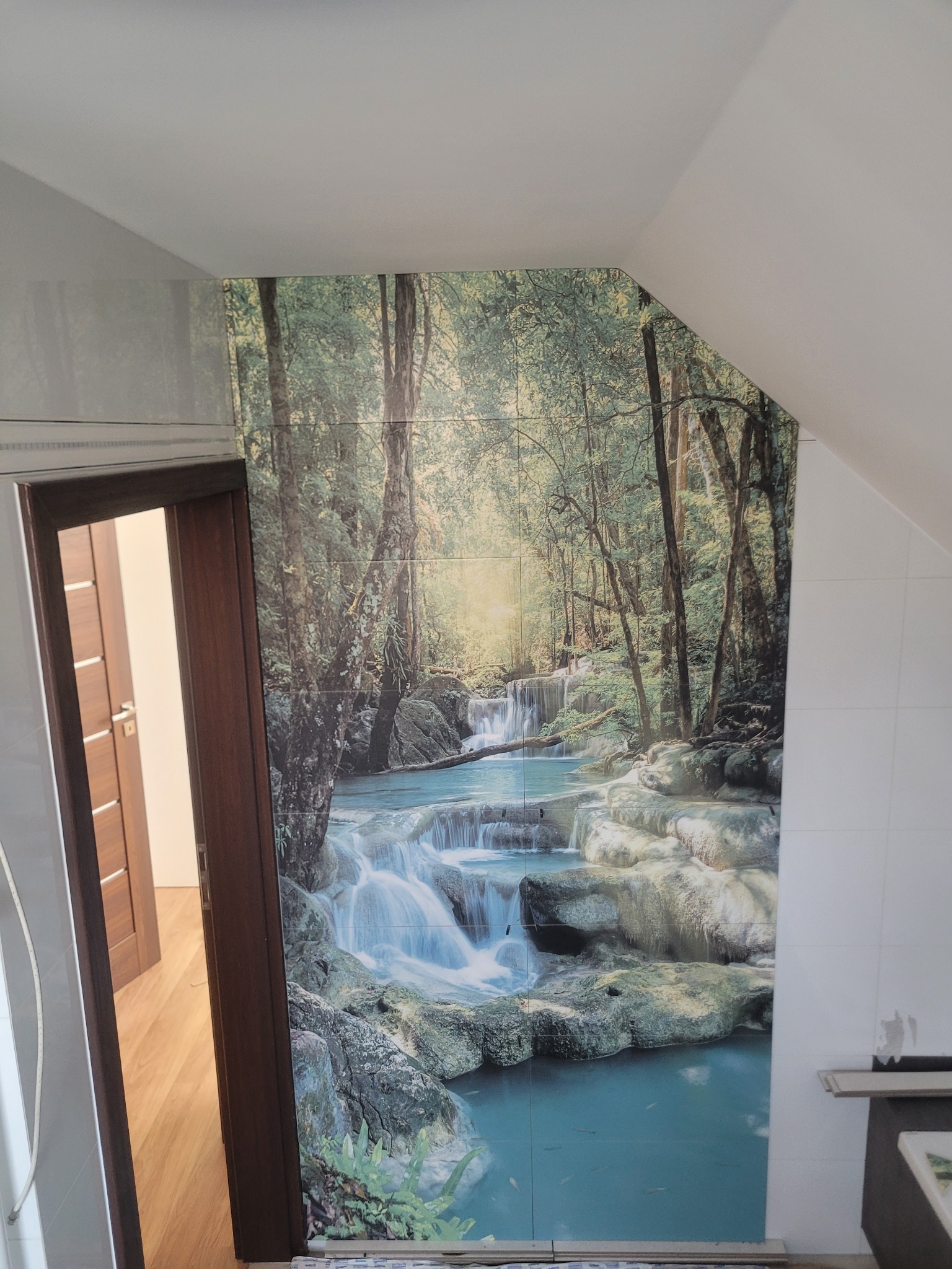 Ściana w łazience wyłożona płytkami z motywem wodospadu w lesie, tworząc iluzję głębi. Widoczny fragment drzwi i skośny sufit.