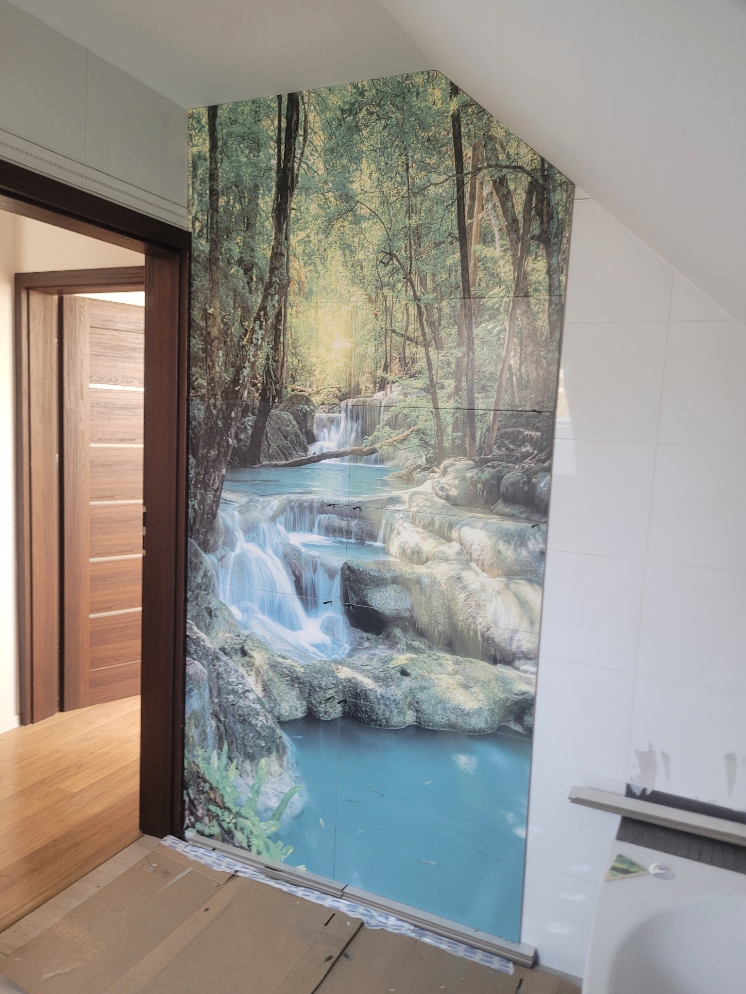 Ściana w łazience wyłożona płytkami z motywem wodospadu w lesie, sąsiadująca z białą ścianą z płytek i wejściem do pokoju z drewnianą podłogą.