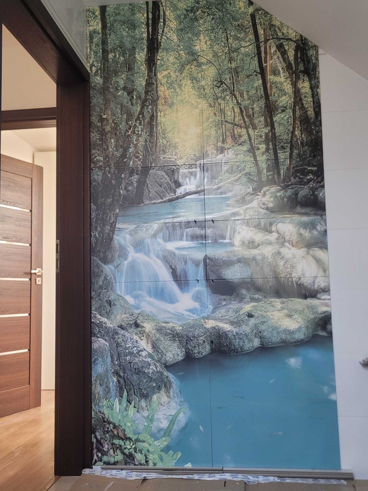 Ściana wyłożona płytkami z motywem wodospadu w lesie, widoczna z perspektywy otwartych drzwi. Płytki tworzą spójny obraz, imitujący fototapetę. Widoczne fugi.