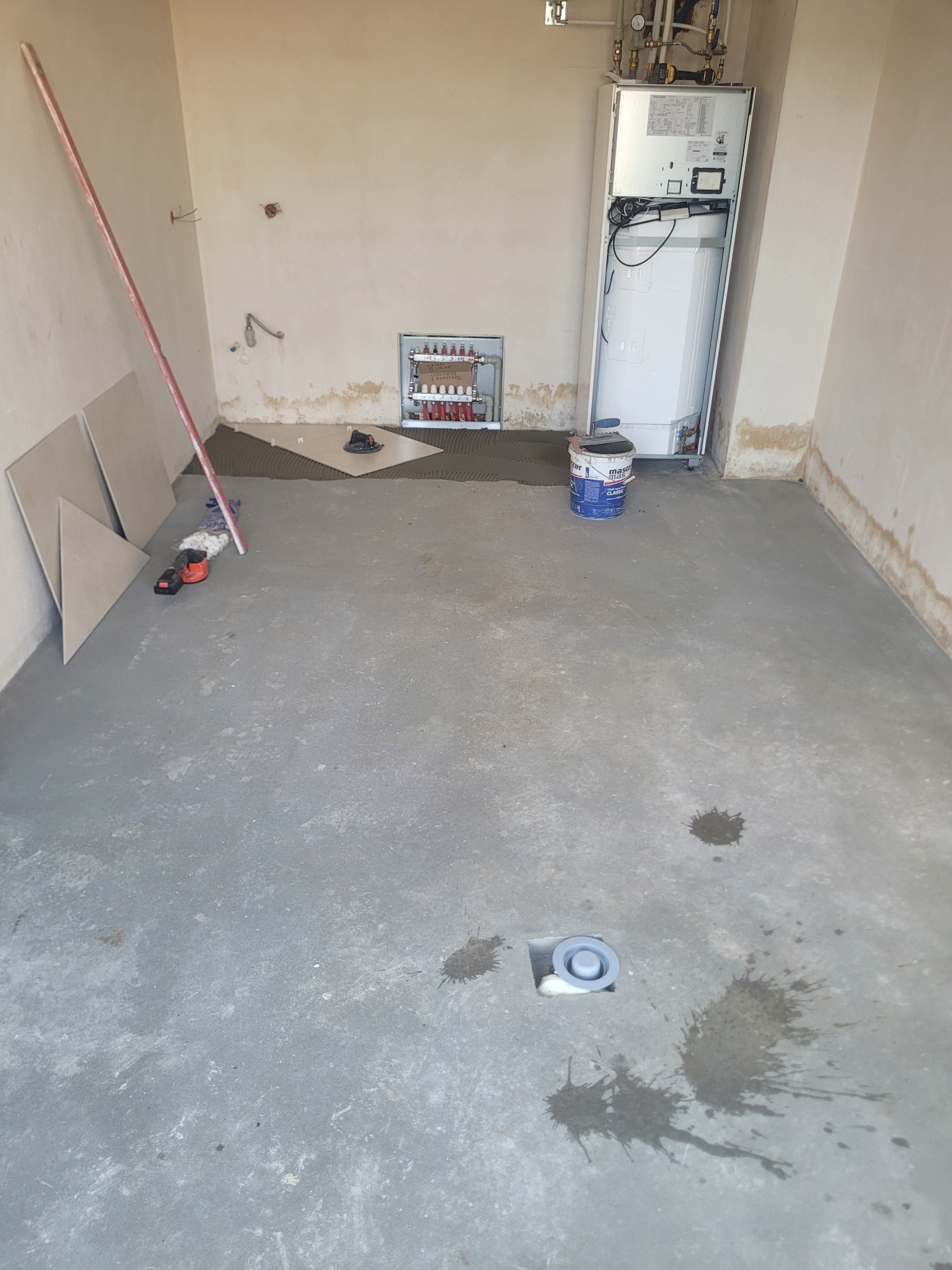 Przygotowanie do układania płytek w łazience: widoczny odpływ liniowy w posadzce, grzejnik, płytki i narzędzia. Etap prac wykończeniowych, surowe ściany.