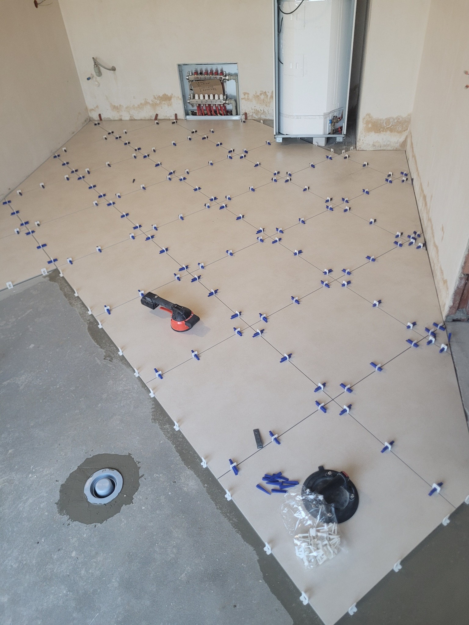 Układanie jasnych płytek podłogowych z użyciem systemu poziomowania. Widoczny fragment surowej posadzki, narzędzia glazurnicze i instalacja CO w tle.