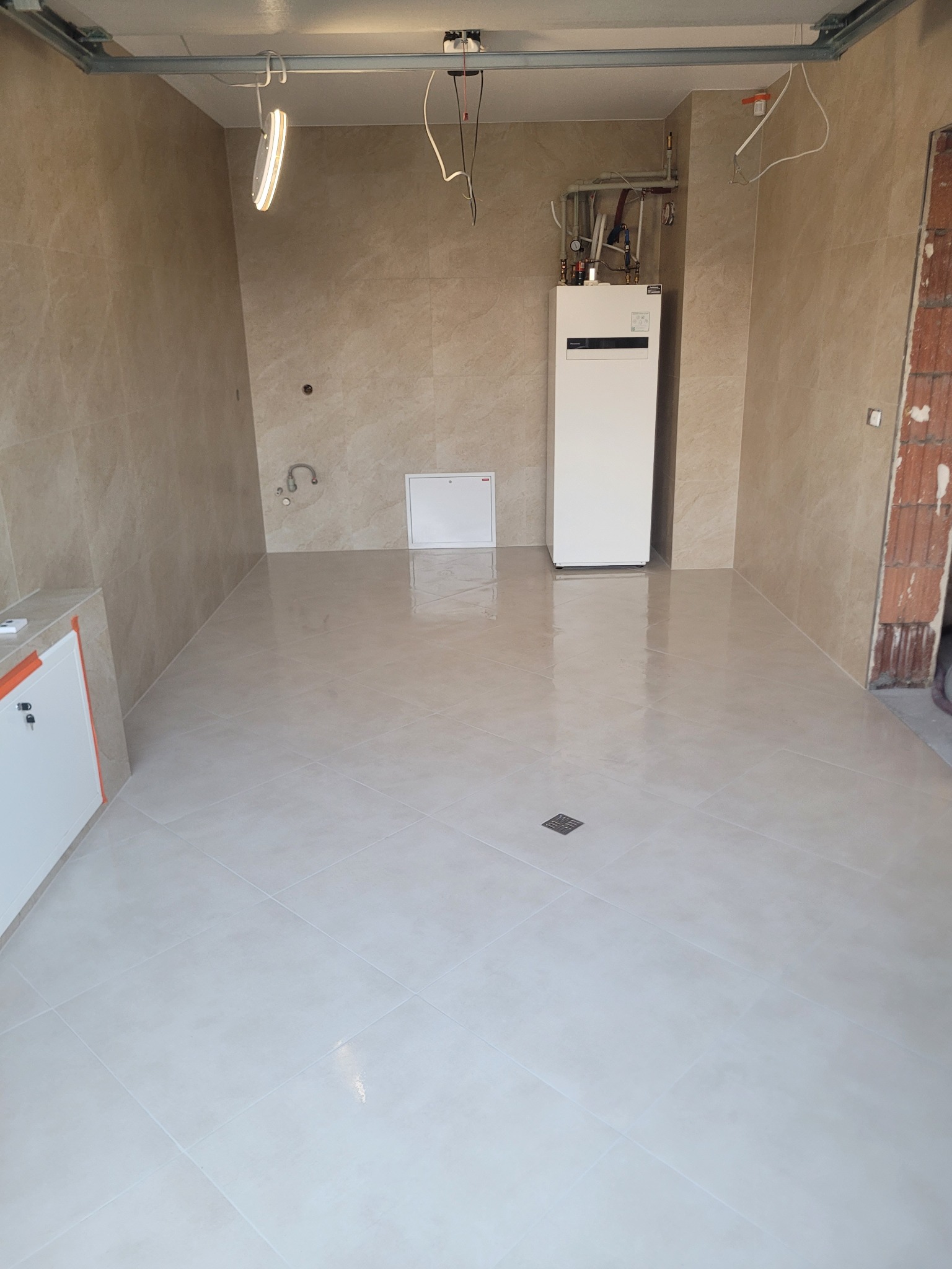 Wykończona łazienka w beżowych płytkach z białym piecem grzewczym i szafką. Widoczny odpływ liniowy w podłodze i surowe wykończenie otworu drzwiowego.