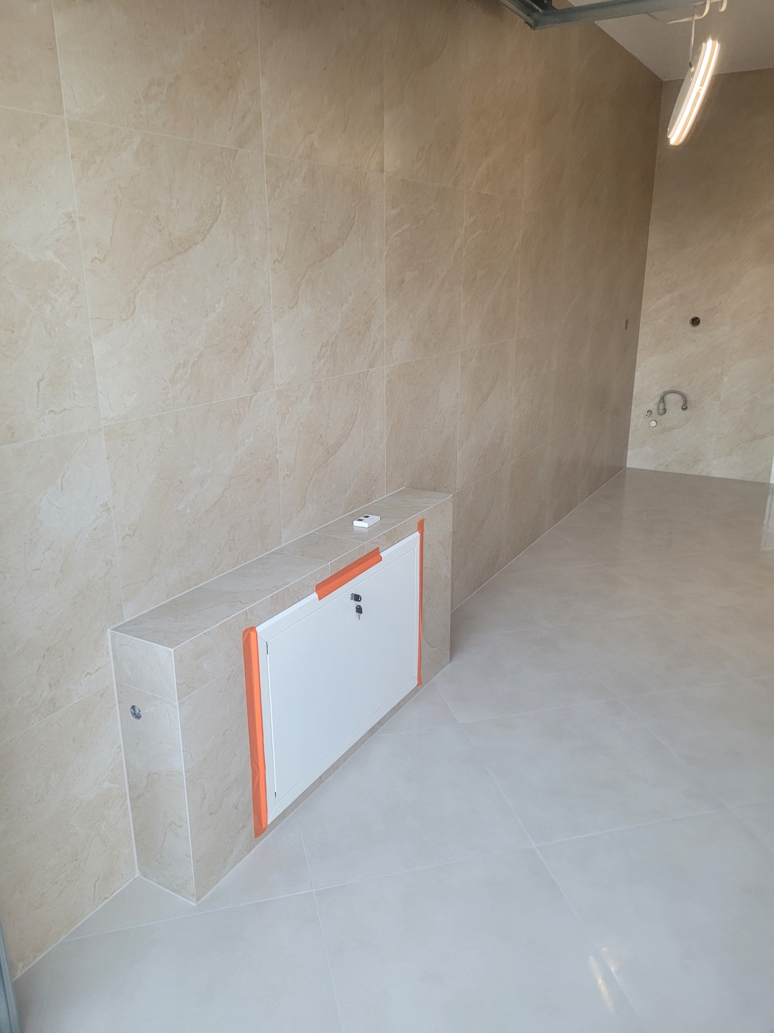 Wykończona łazienka z beżowymi płytkami imitującymi marmur na ścianach i podłodze. Widoczna zabudowa z drzwiczkami rewizyjnymi, zabezpieczona pomarańczową taśmą.