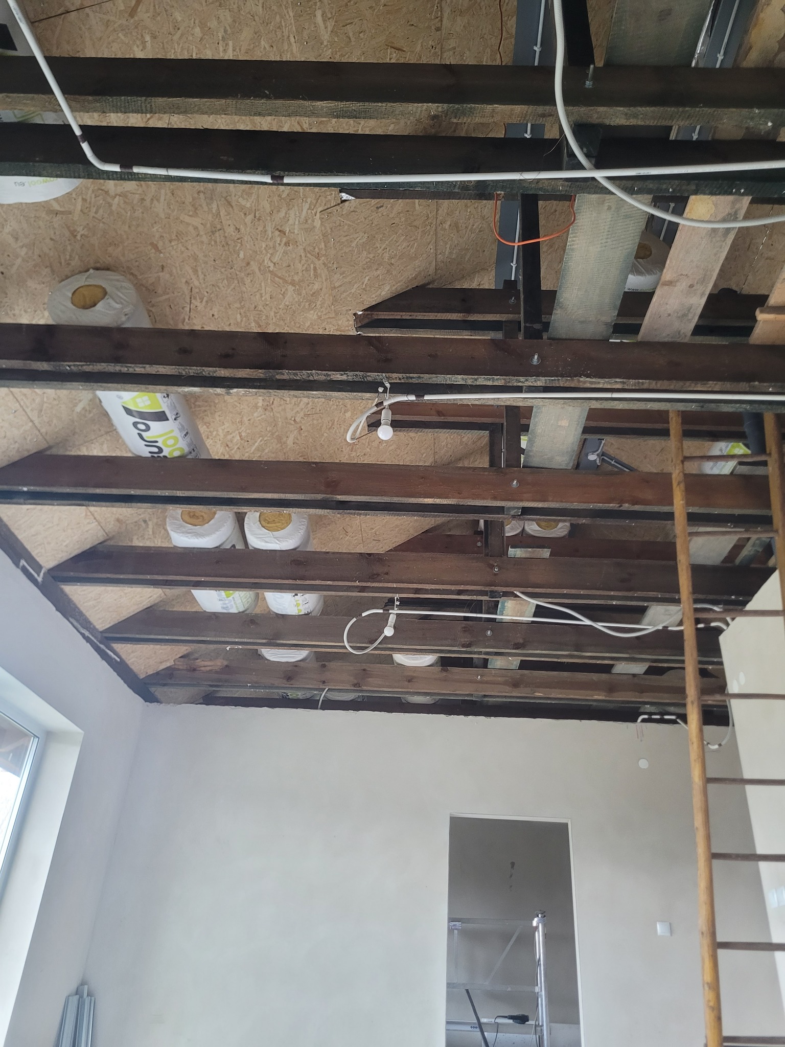 Poddasze w trakcie ocieplania: widoczne drewniane belki, wełna mineralna, instalacja elektryczna i fragment ściany z oknem. Etap prac przed wykończeniem.