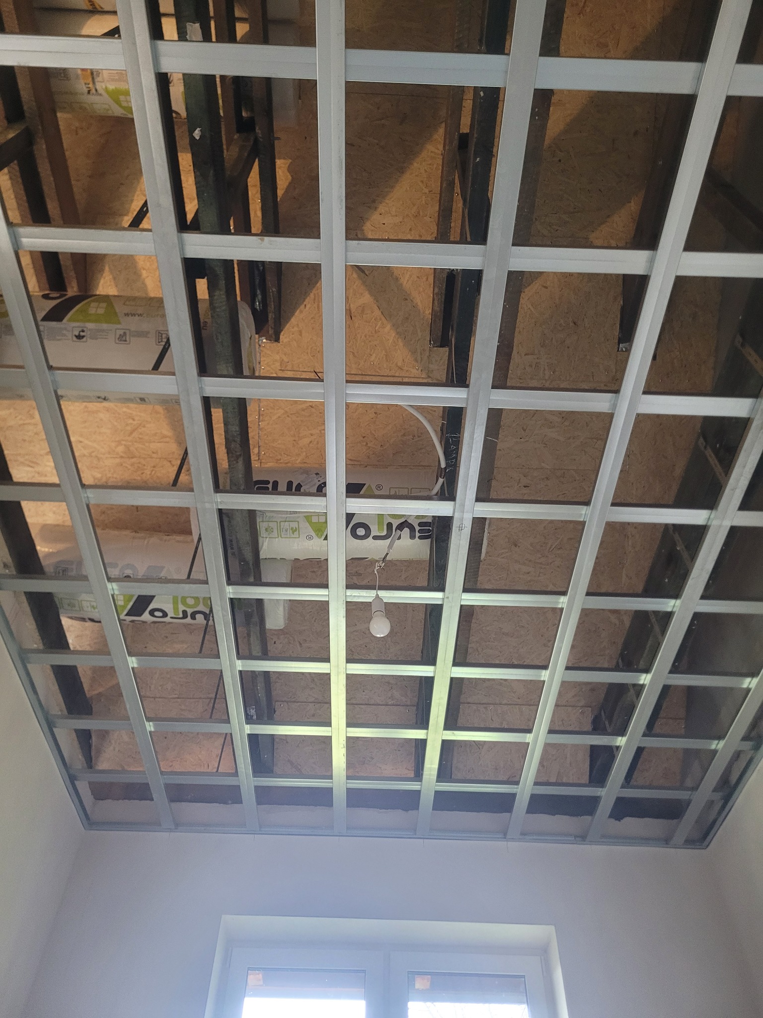 Metalowa konstrukcja sufitu podwieszanego na poddaszu, widoczne ocieplenie, surowe drewno i instalacja elektryczna z żarówką. Ujęcie z dołu, widoczne okno.