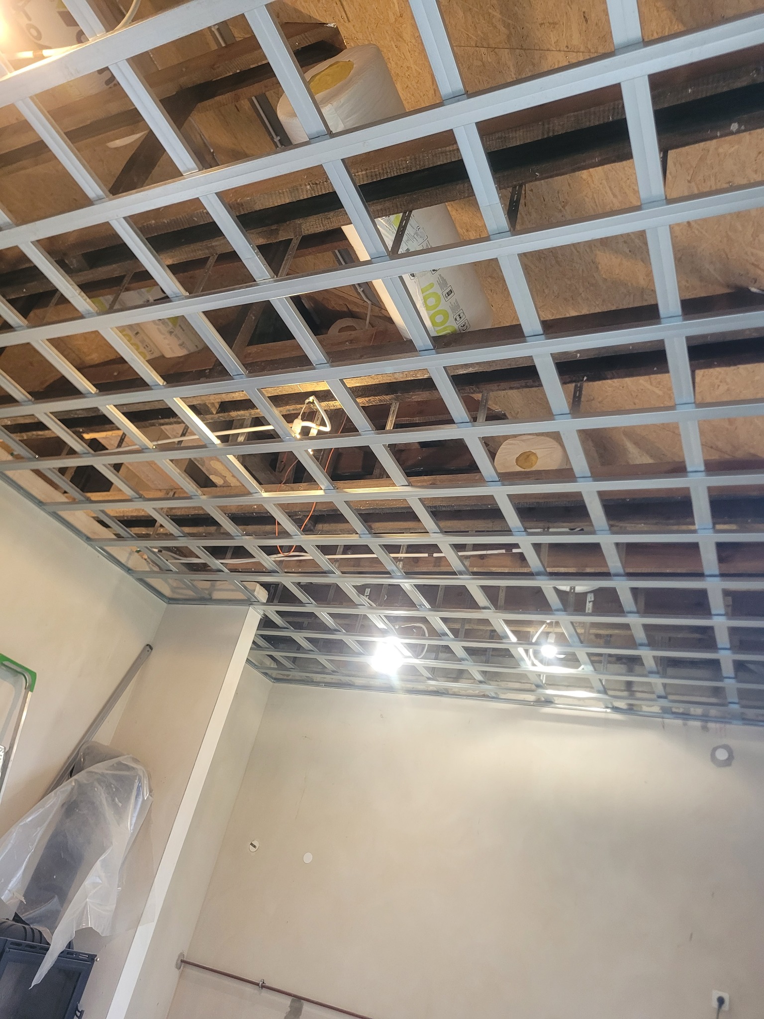 Sufit podwieszany z metalową konstrukcją kratową, widoczne ocieplenie poddasza i elementy instalacji. Prace wykończeniowe w trakcie realizacji.