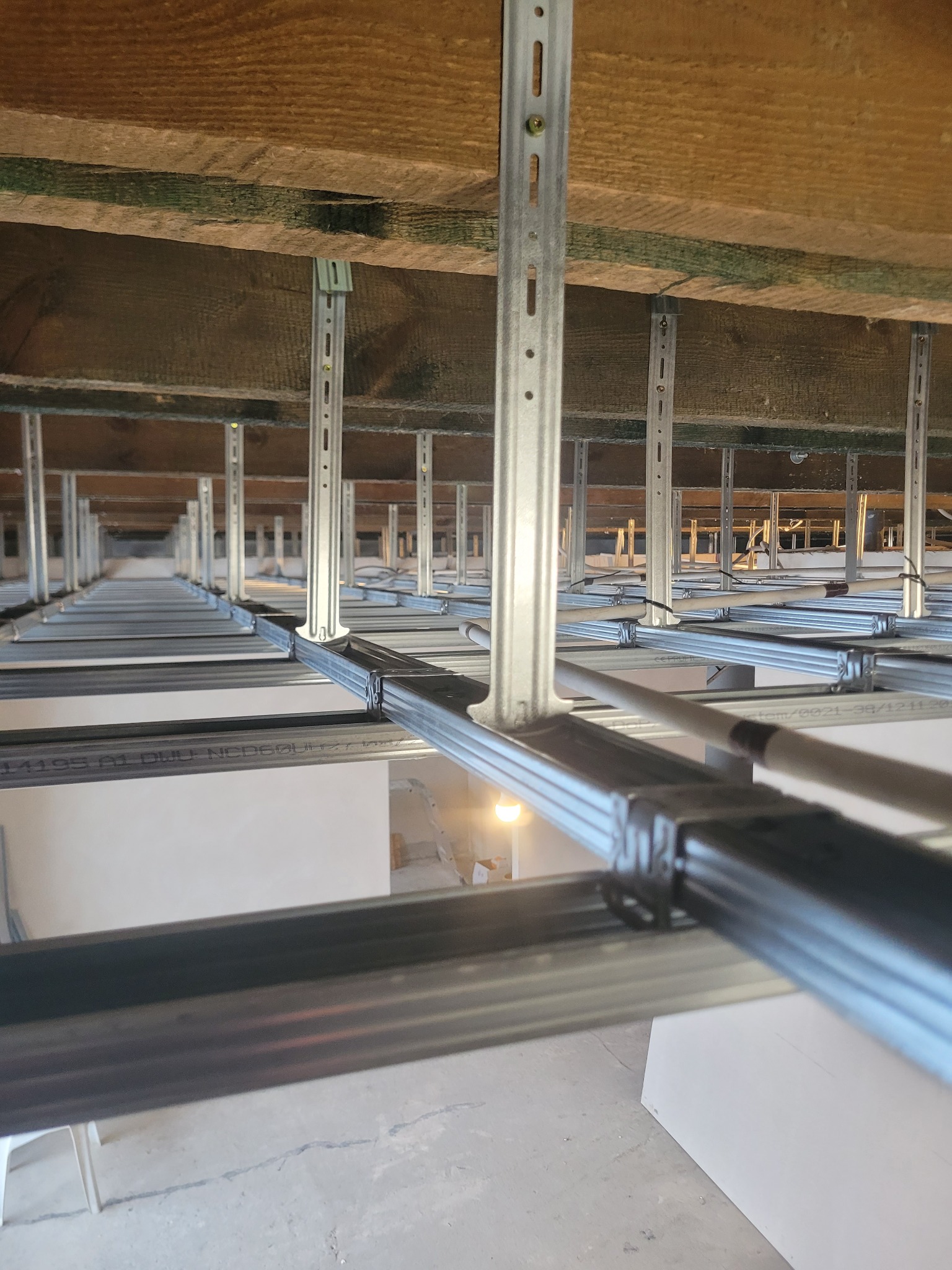 Metalowa konstrukcja podwieszanego sufitu z płyt karton-gips, widok z dołu, z perspektywą zbieżną w głąb pomieszczenia. Elementy montażowe widoczne na drewnianym stropie.