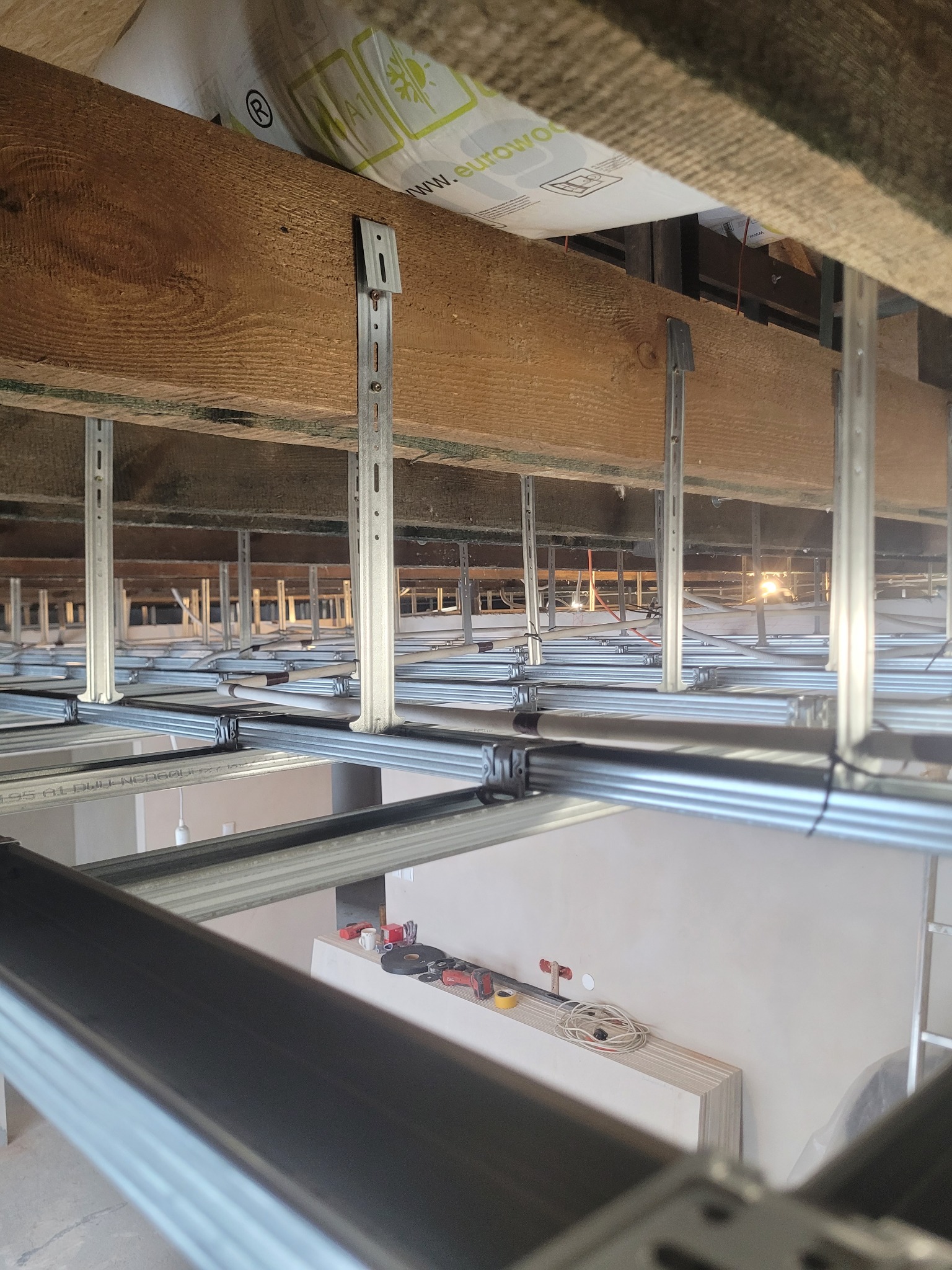 Stelaż sufitu podwieszanego z profili metalowych, zamocowany do drewnianej konstrukcji stropu. Widoczne narzędzia i materiały budowlane w tle.