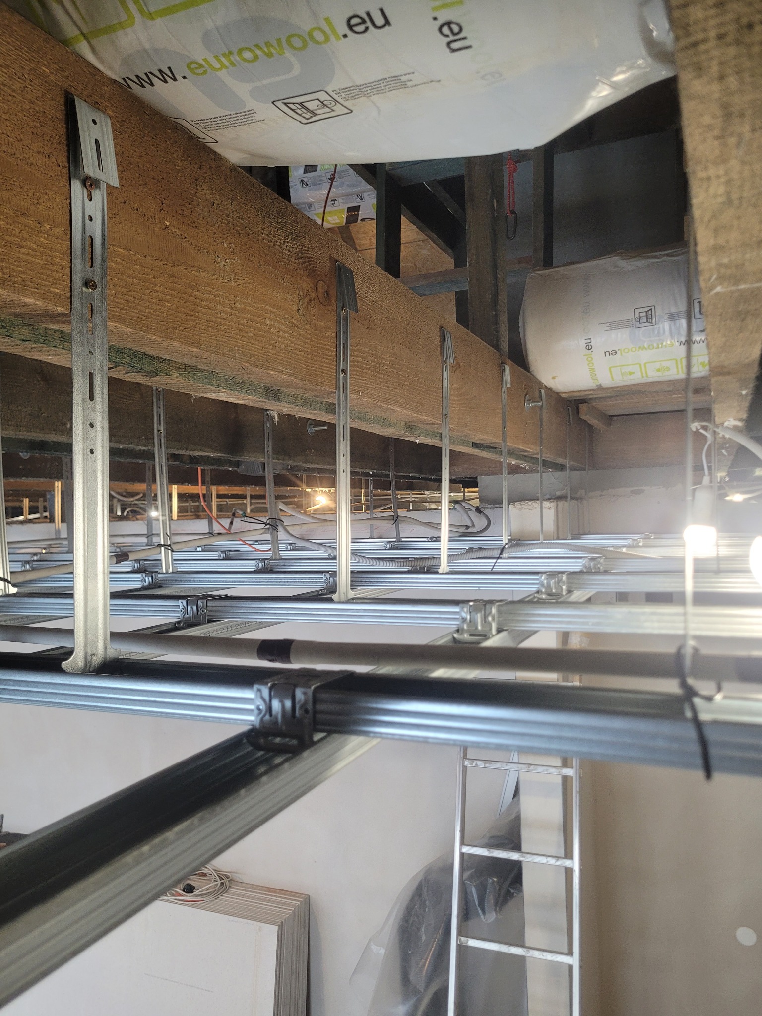 Sufit podwieszany w trakcie montażu: metalowa konstrukcja z profilami, widoczne elementy mocujące do drewnianych belek, ocieplenie z wełny mineralnej. Perspektywa z dołu.