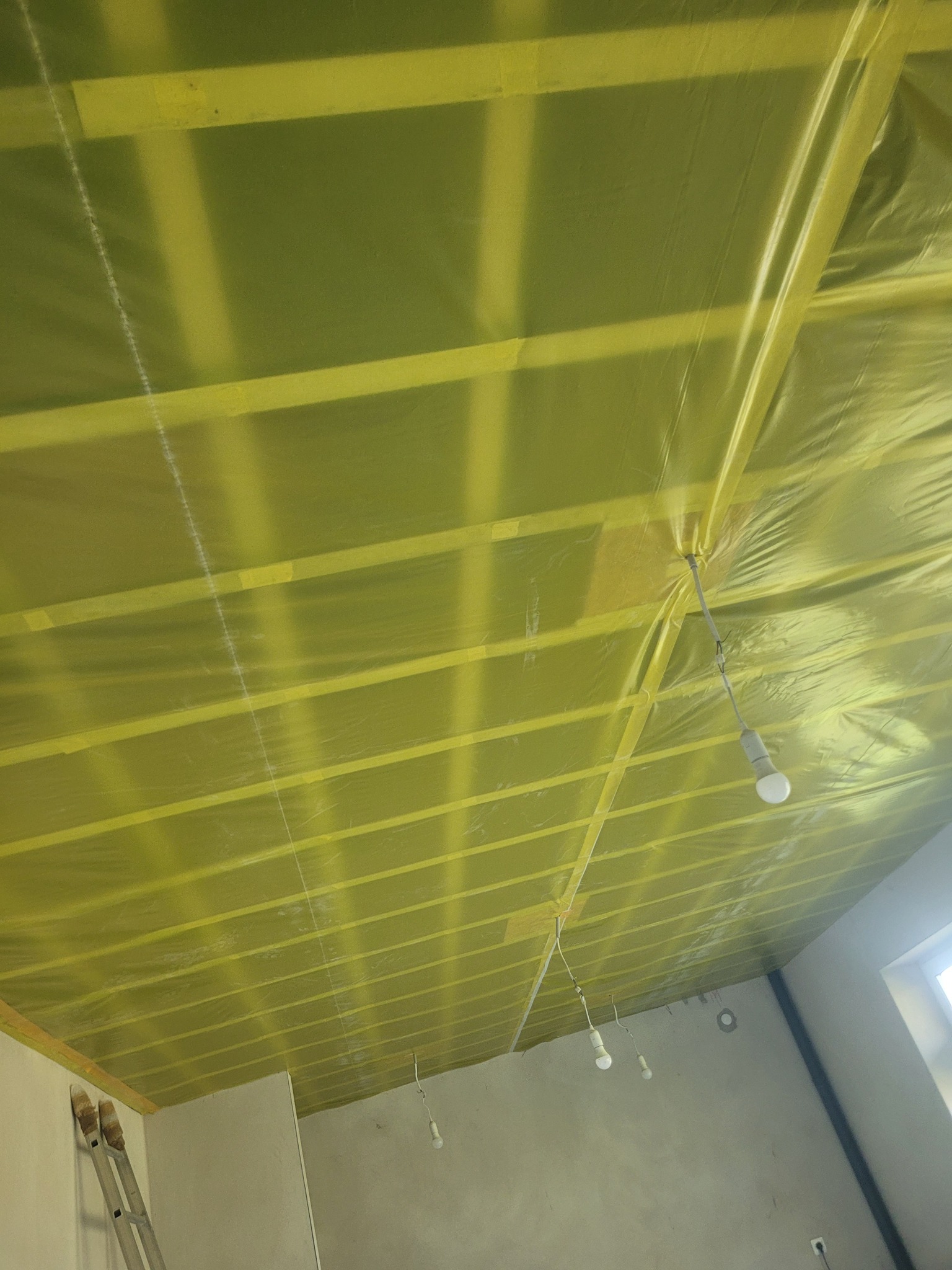 Żółta folia paroizolacyjna na drewnianej konstrukcji poddasza, widok z dołu, drabina oparta o ścianę, surowy stan wykończenia wnętrza.